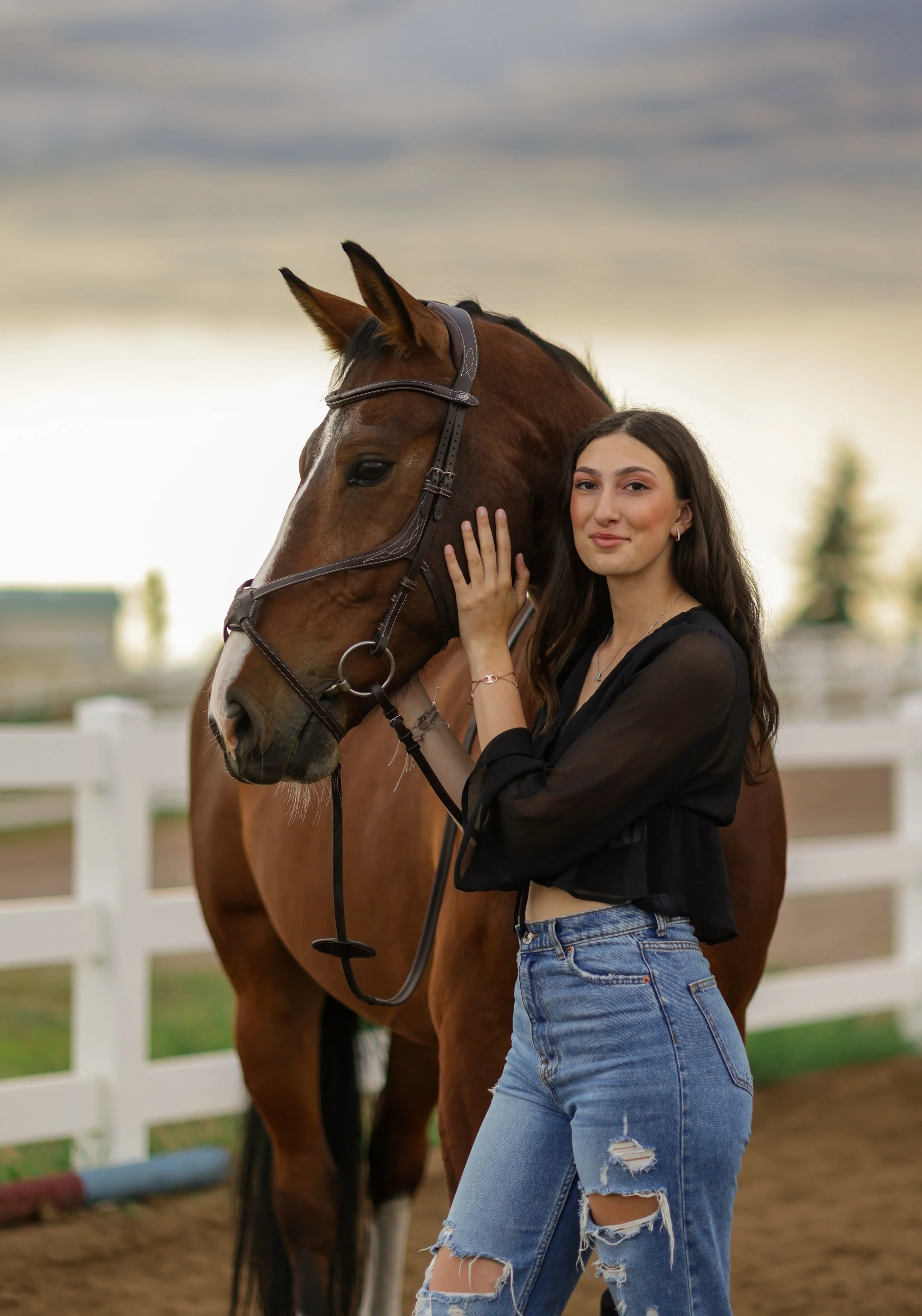 A young woman standing next to a brown horse with a white stripe on its face, holding its bridle and kissing its neck, at an outdoor equestrian area with a white fence and cloudy sky in the background.