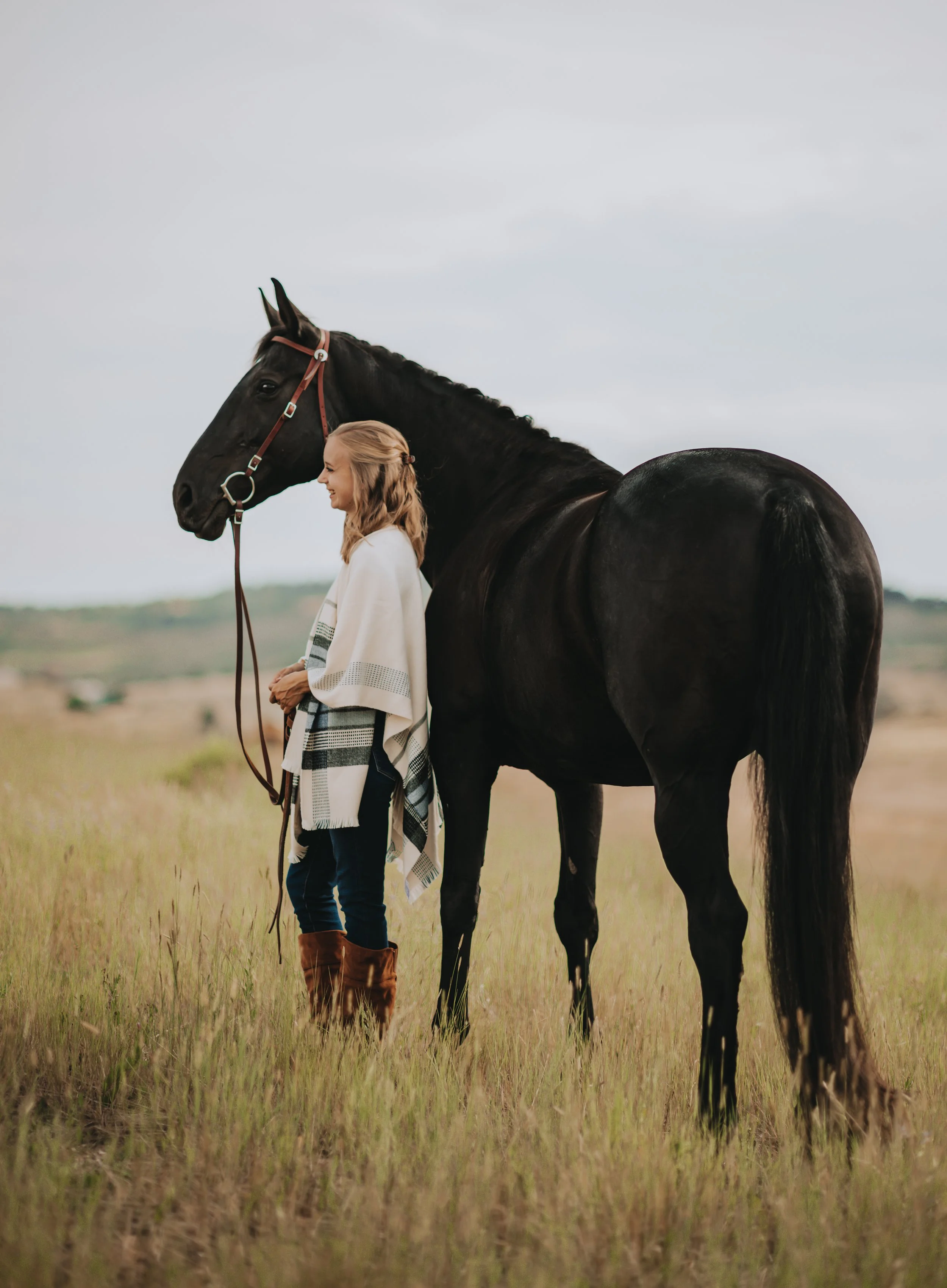 A woman smiling and wearing a white shawl stands in a grassy field next to a tall black horse with a brown harness, under a cloudy sky.