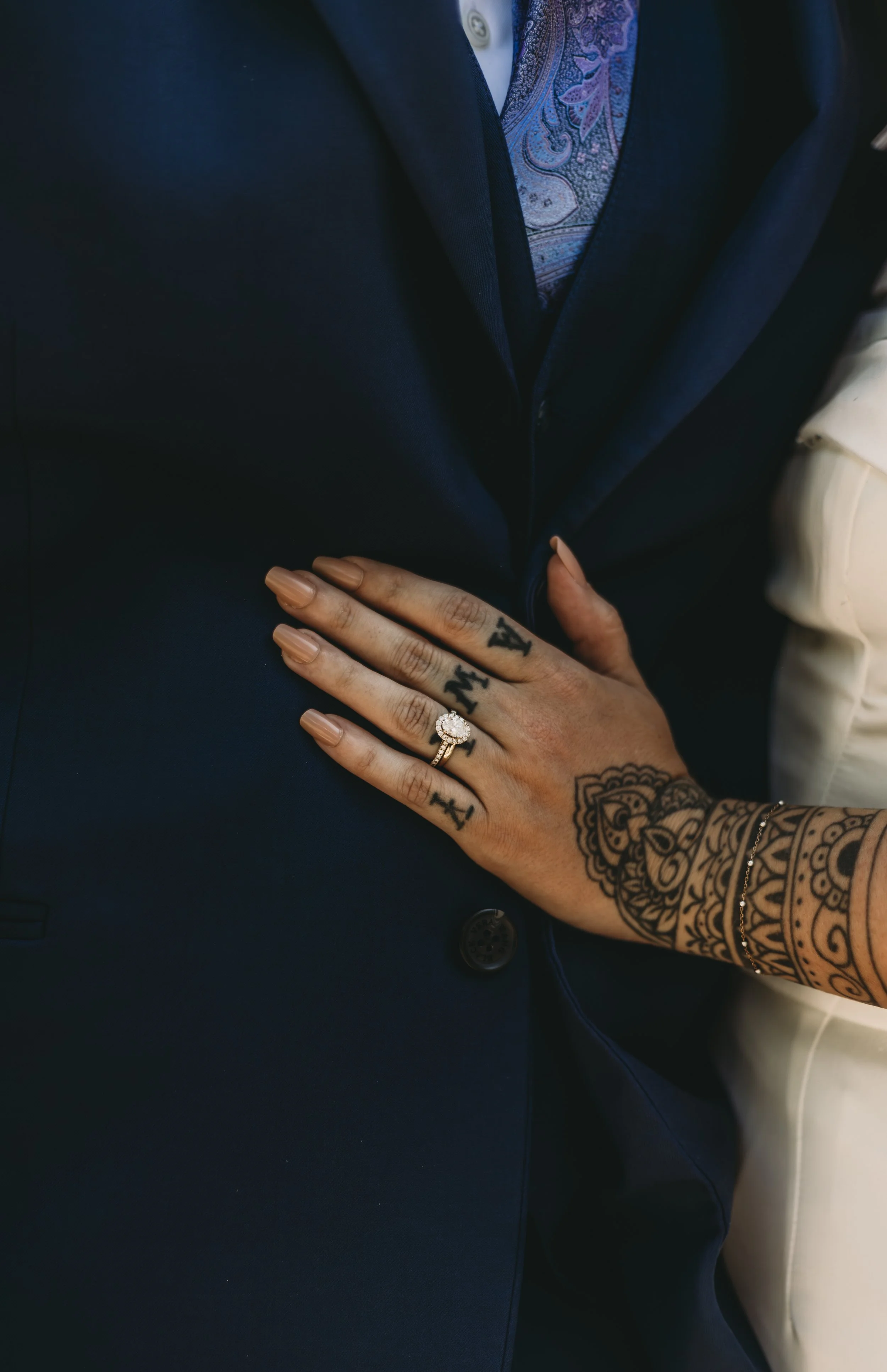 Close-up of a person's hand with tattoos, showing a ring with a large gemstone, resting on a dark suit near a patterned shirt.