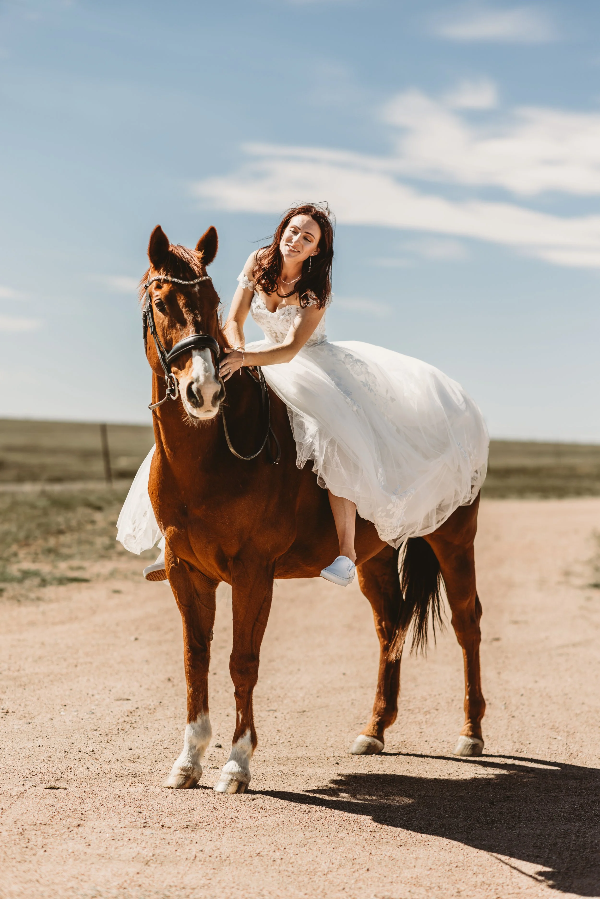A woman in a wedding dress riding a chestnut horse on a dirt path in a rural area with open fields and a partly cloudy sky.