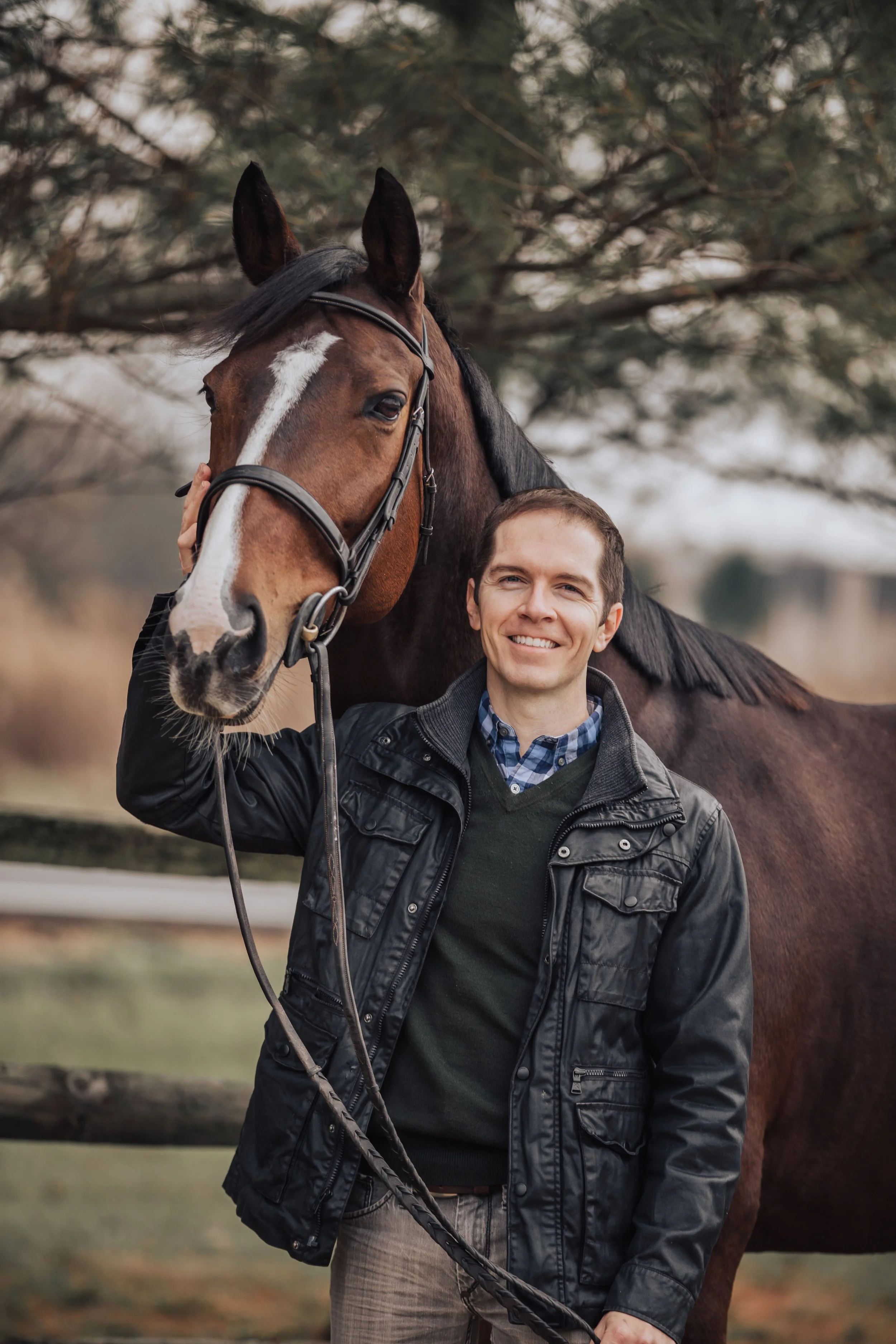 A man in a black jacket smiling and holding a horse's bridle, outdoors with trees in the background.