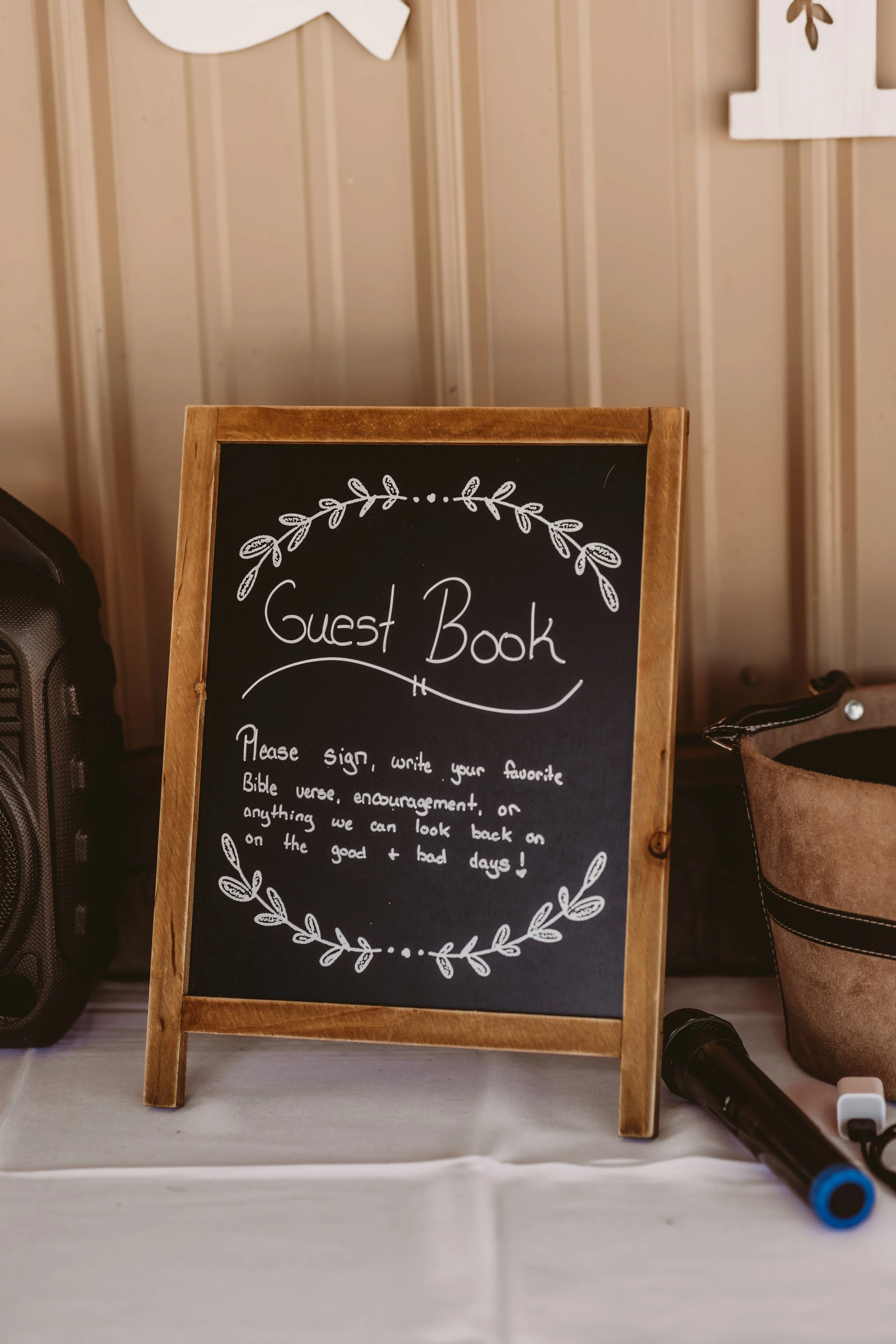A black chalkboard sign with a wooden frame displaying a message about a guest book, surrounded by white decorative leaf illustrations, placed on a table with a marker nearby.
