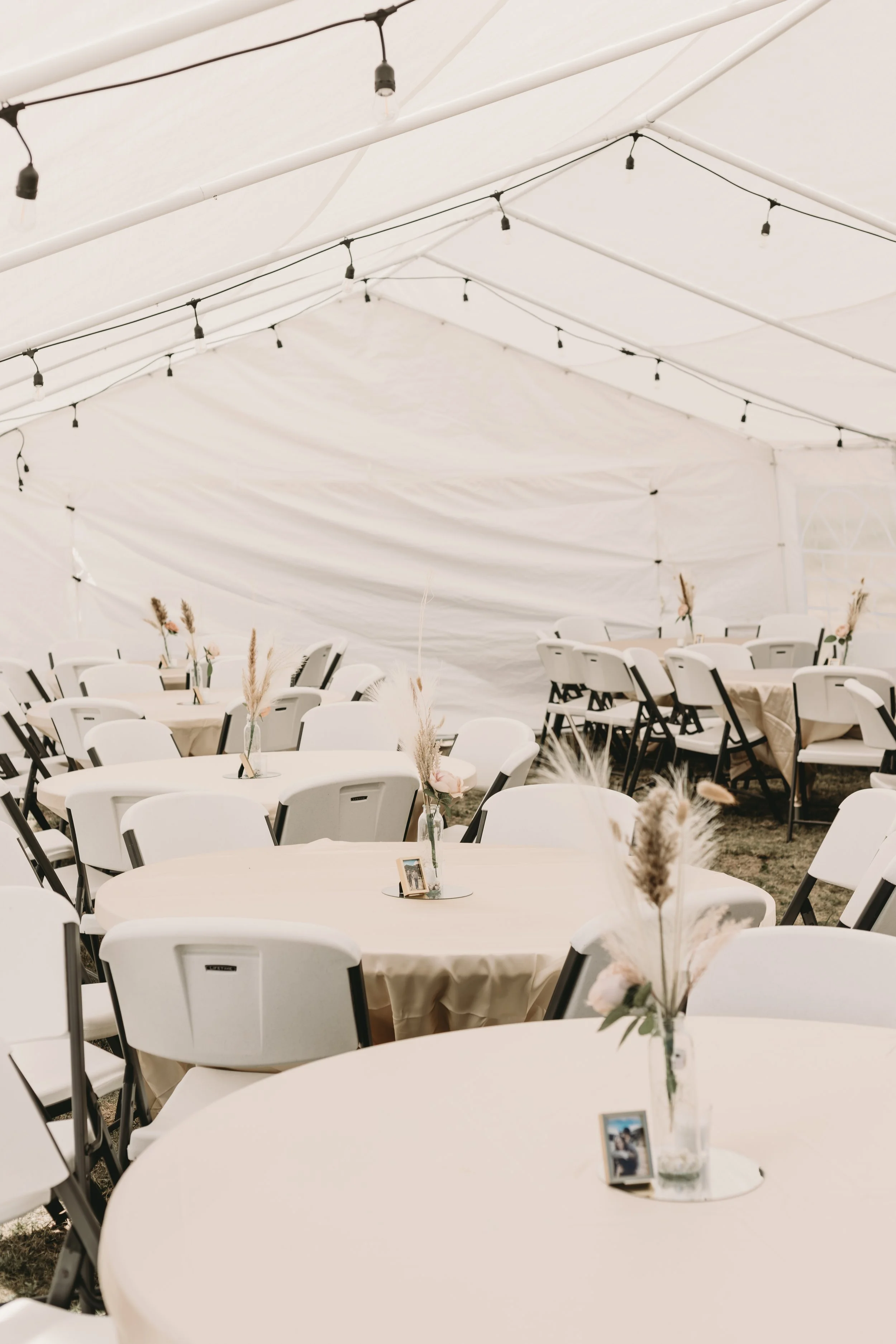 White event tent with string lights, decorated tables with centerpieces, and white chairs for a celebration or gathering.