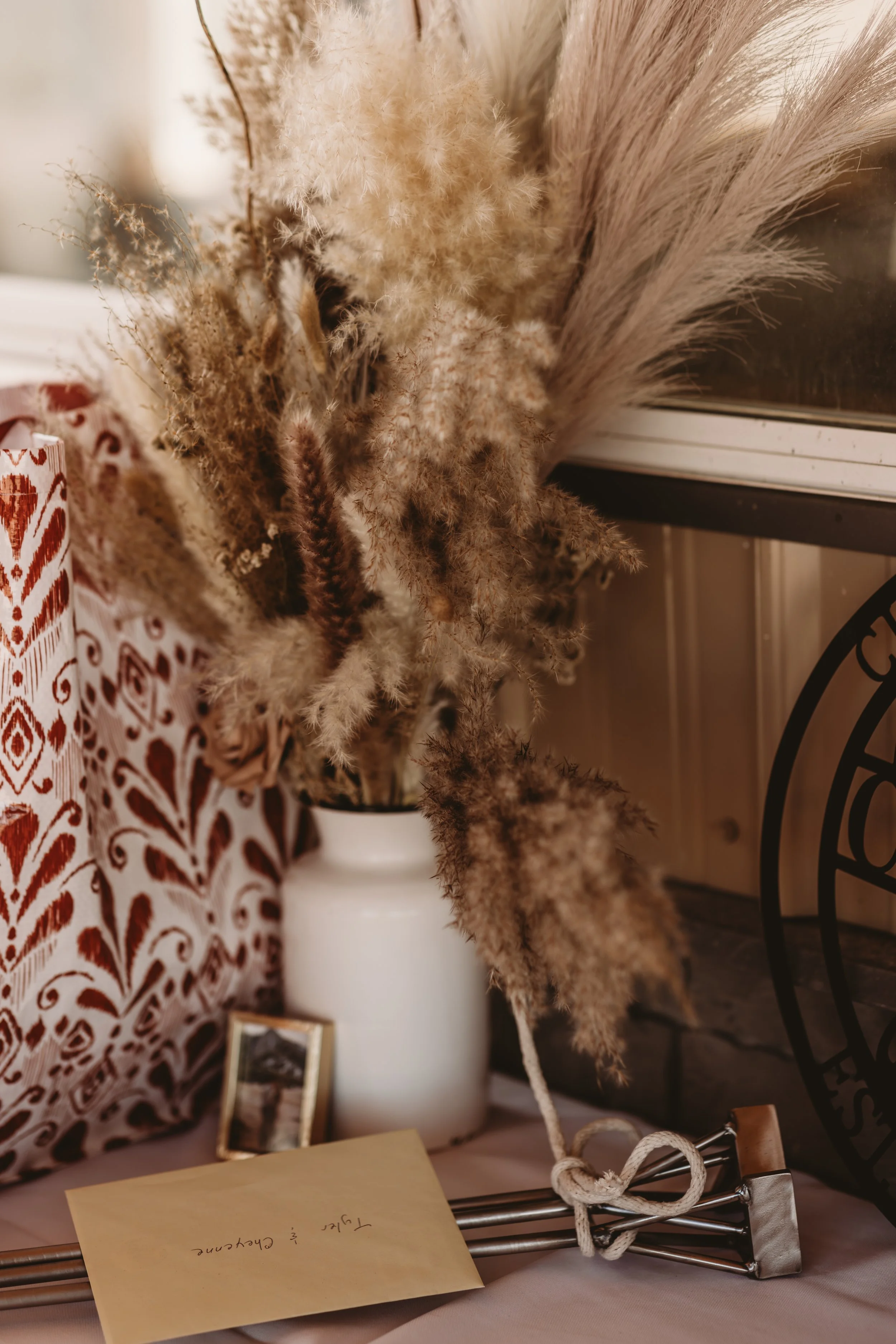 A white vase with dried beige and brown pampas grass and fluffy seed heads, placed on a table next to a patterned red and white pillow and a small framed photo, with a note that says 'Tian, & Cheyenne' in front of the vase.