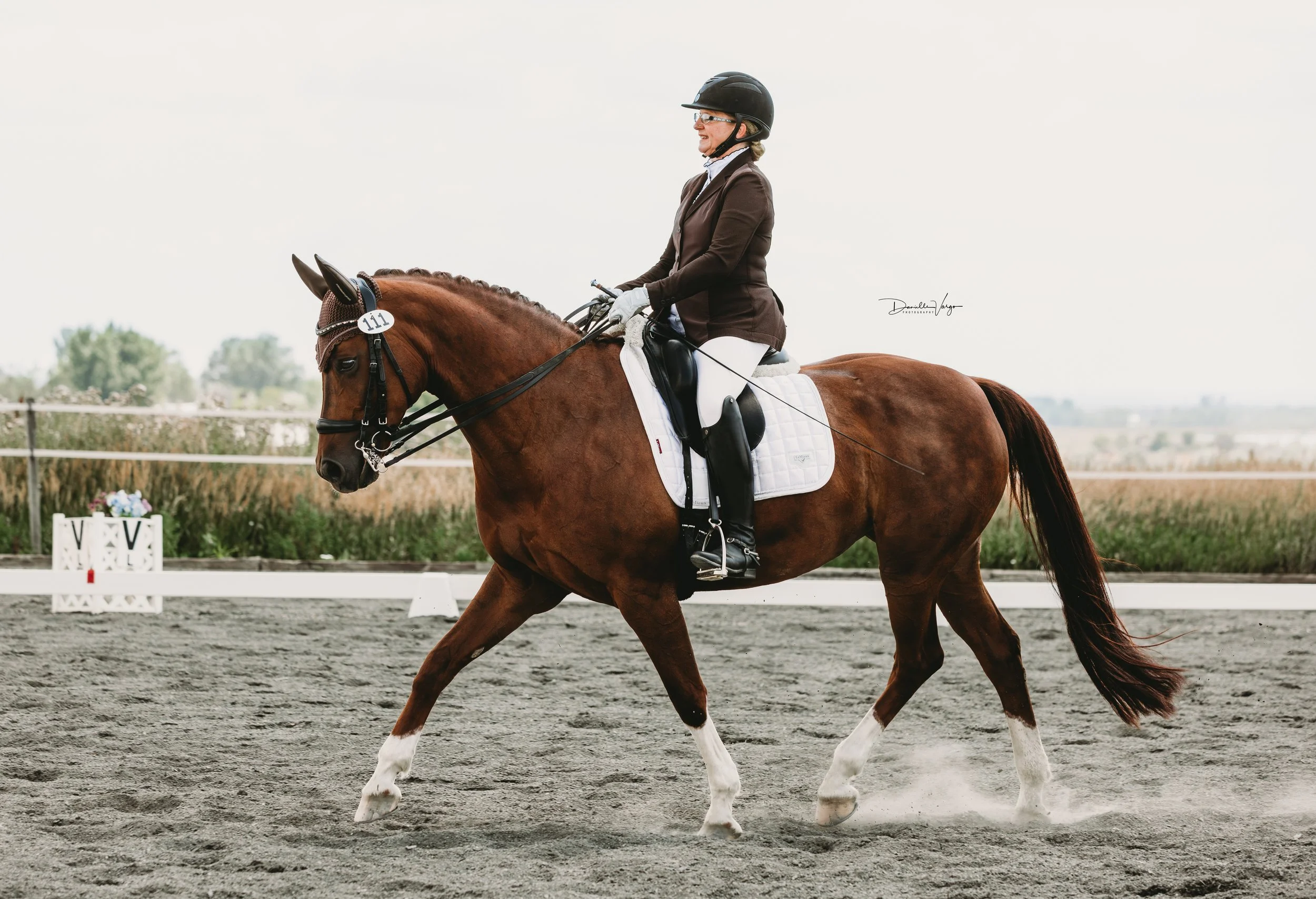 Woman dressed in equestrian attire riding a brown horse with white markings in an outdoor sand arena.