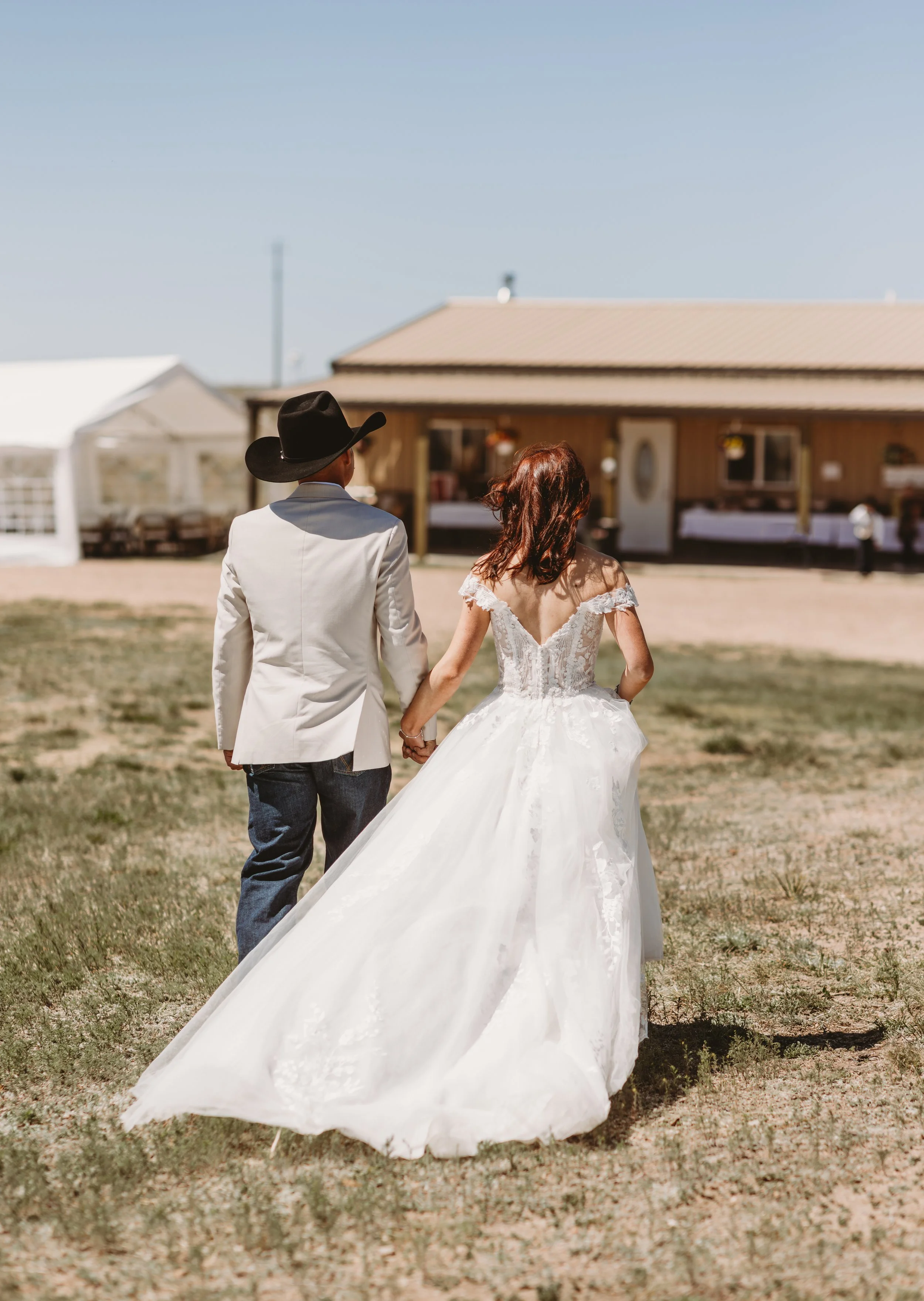 A couple dressed in wedding attire walking hand in hand outdoors on a grassy area with a wooden building in the background.