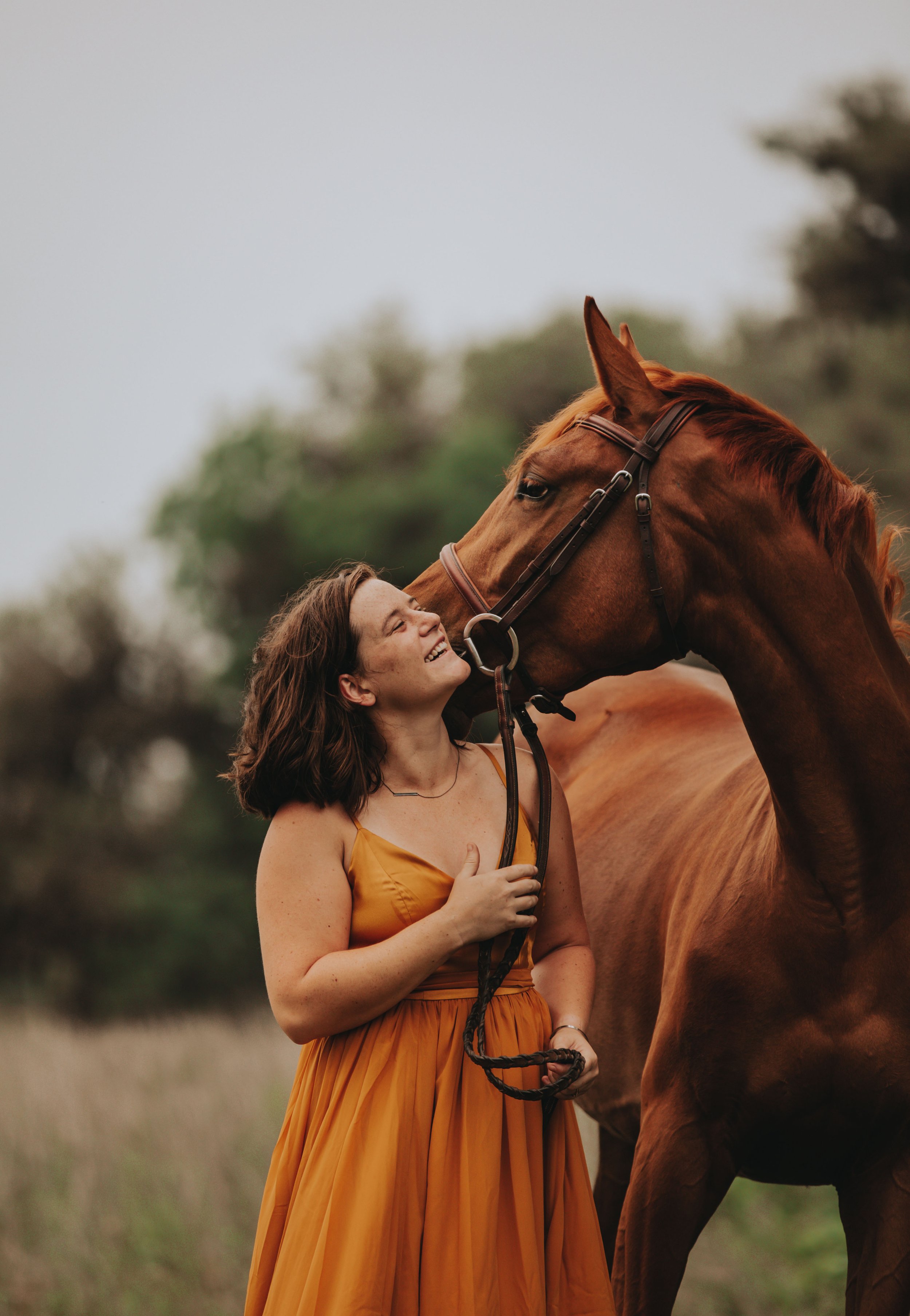 A smiling woman in a mustard-colored dress stands next to a brown horse with a bridle on, outdoors with blurred trees in the background.