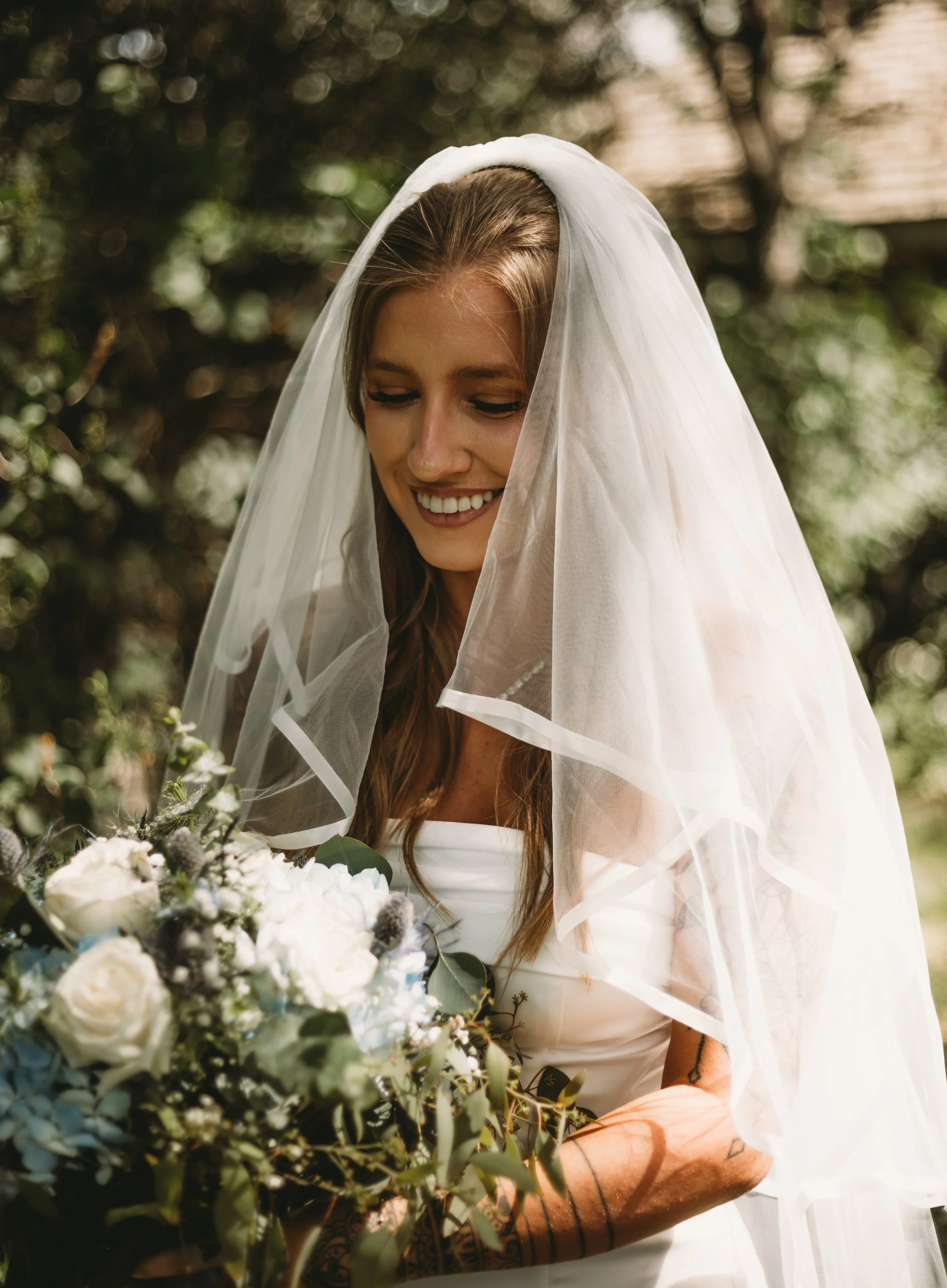 A smiling woman in a wedding dress with a veil, surrounded by flowers outdoors.