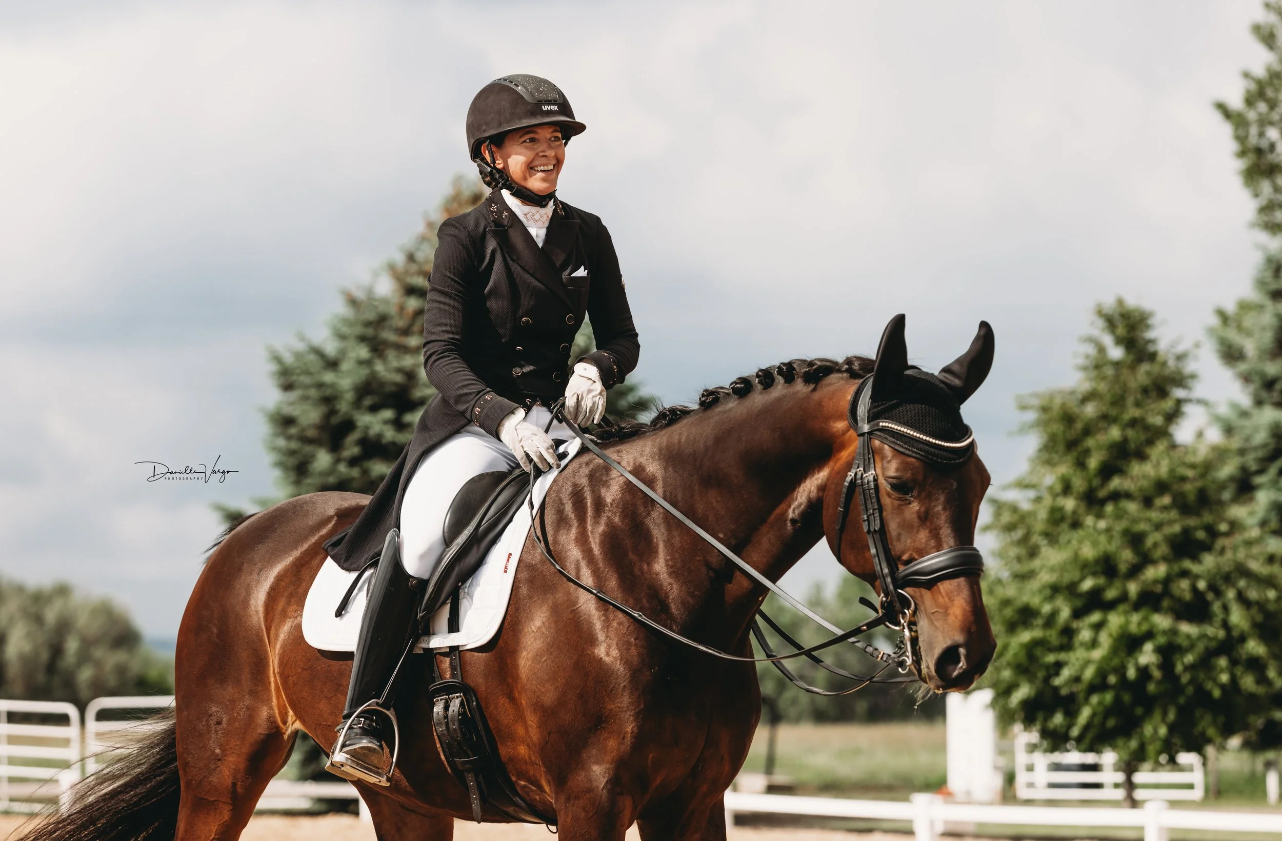 Smiling woman in formal riding attire, wearing a helmet, riding a brown horse outdoors on a cloudy day.