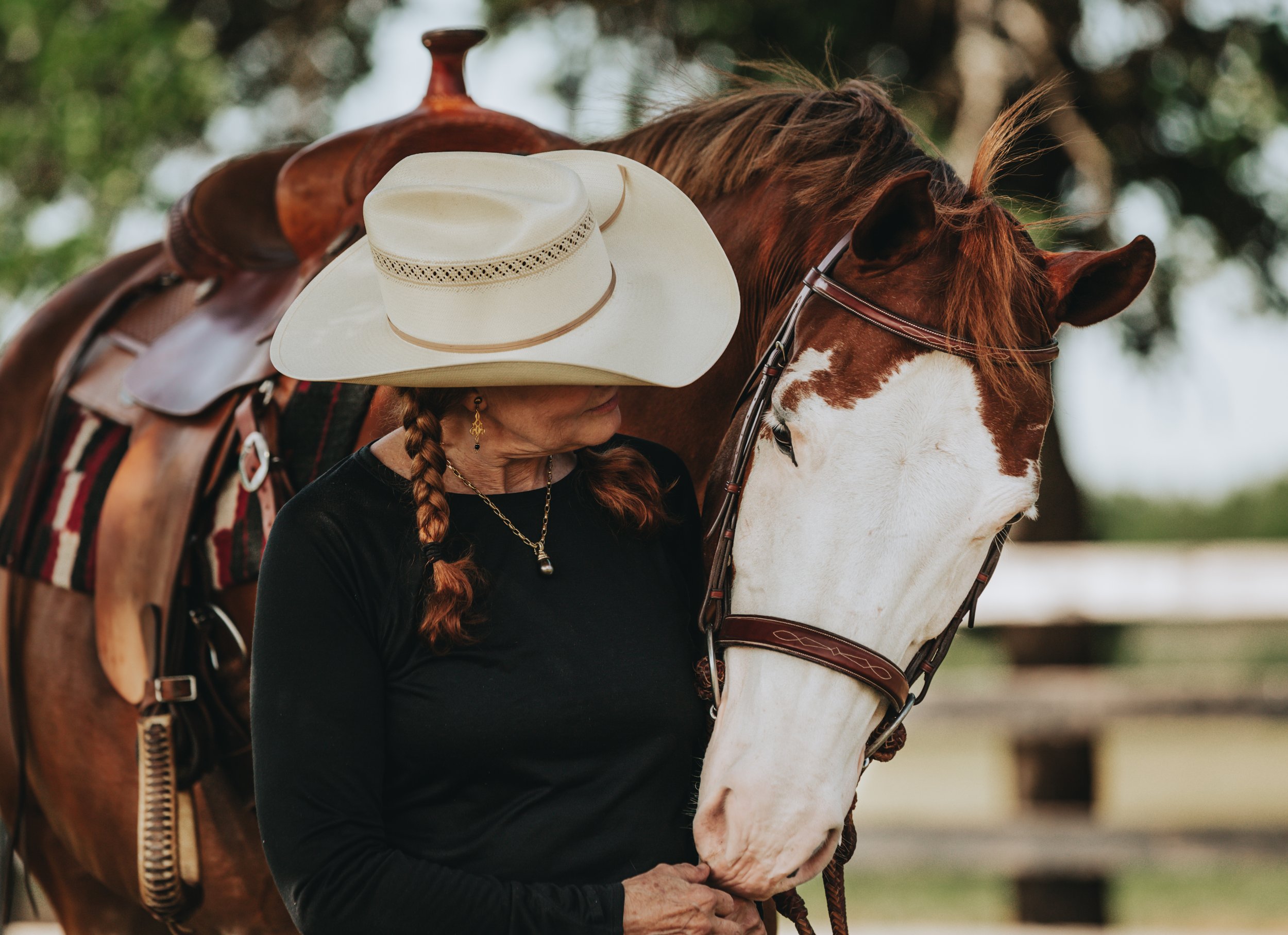 A woman with red hair braided over her shoulder, wearing a black shirt and gold jewelry, is gently holding the muzzle of a white and chestnut horse. The horse is equipped with a leather bridle and saddle, with a white cowboy hat resting on the saddle