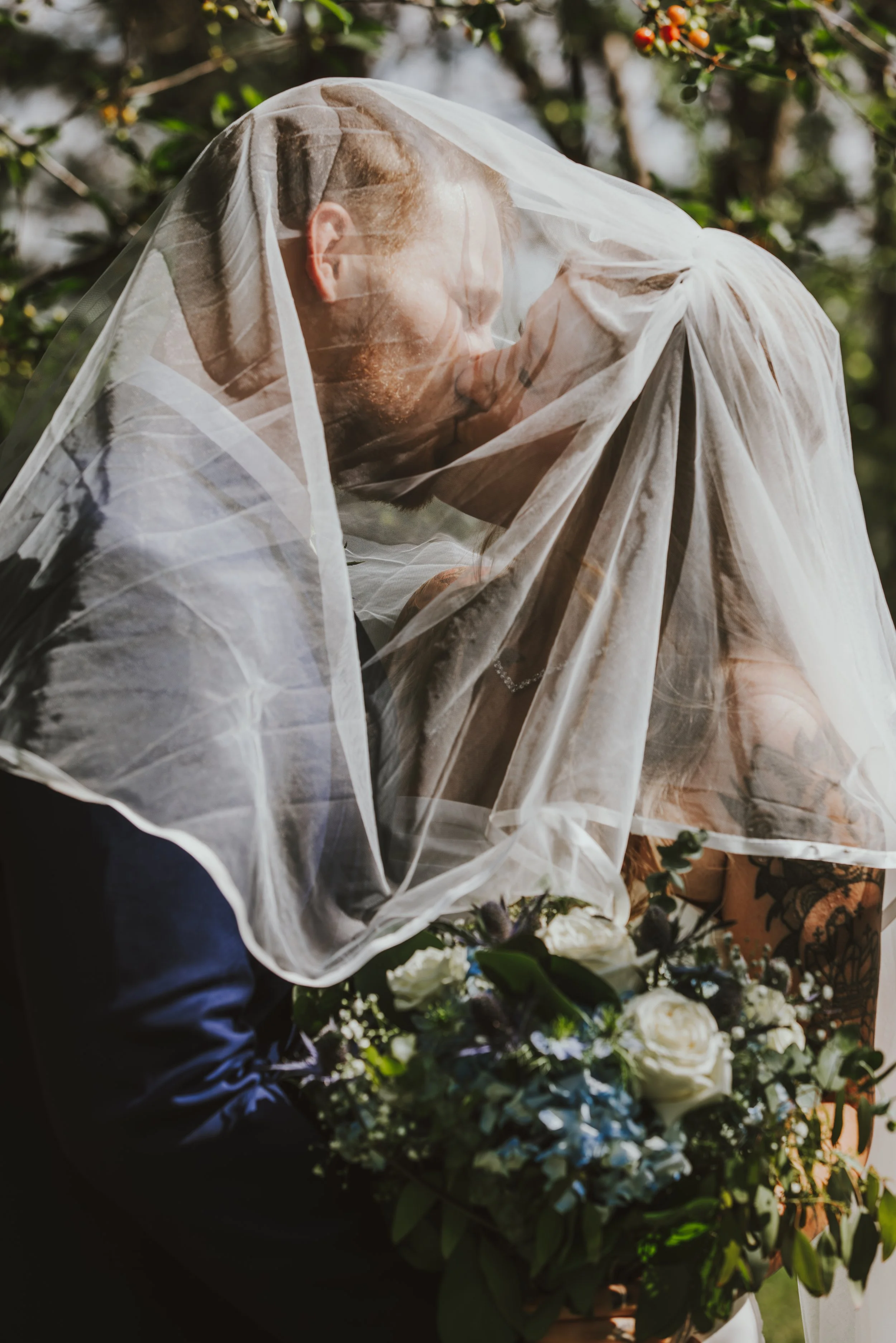 Close-up of a bride and groom kissing under a sheer veil with green foliage and red berries in the background, surrounded by a bouquet of white and green flowers.