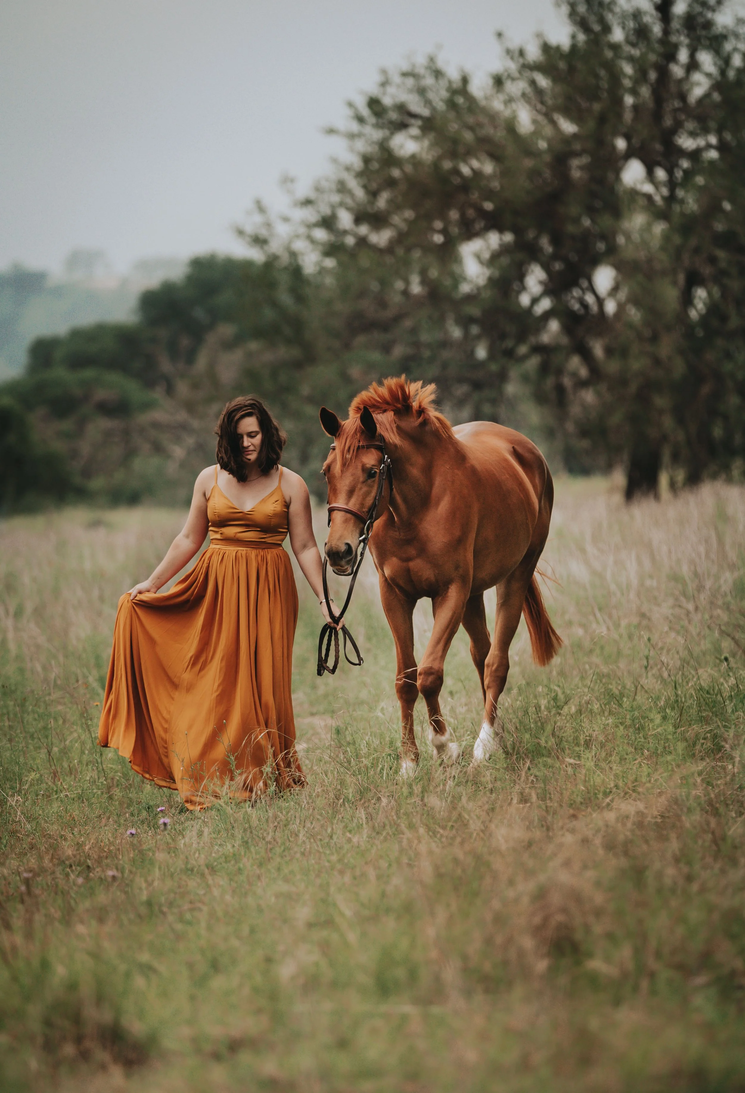 A woman with shoulder-length dark hair wearing a long orange dress, walking in a grassy field while holding the reins of a chestnut horse.