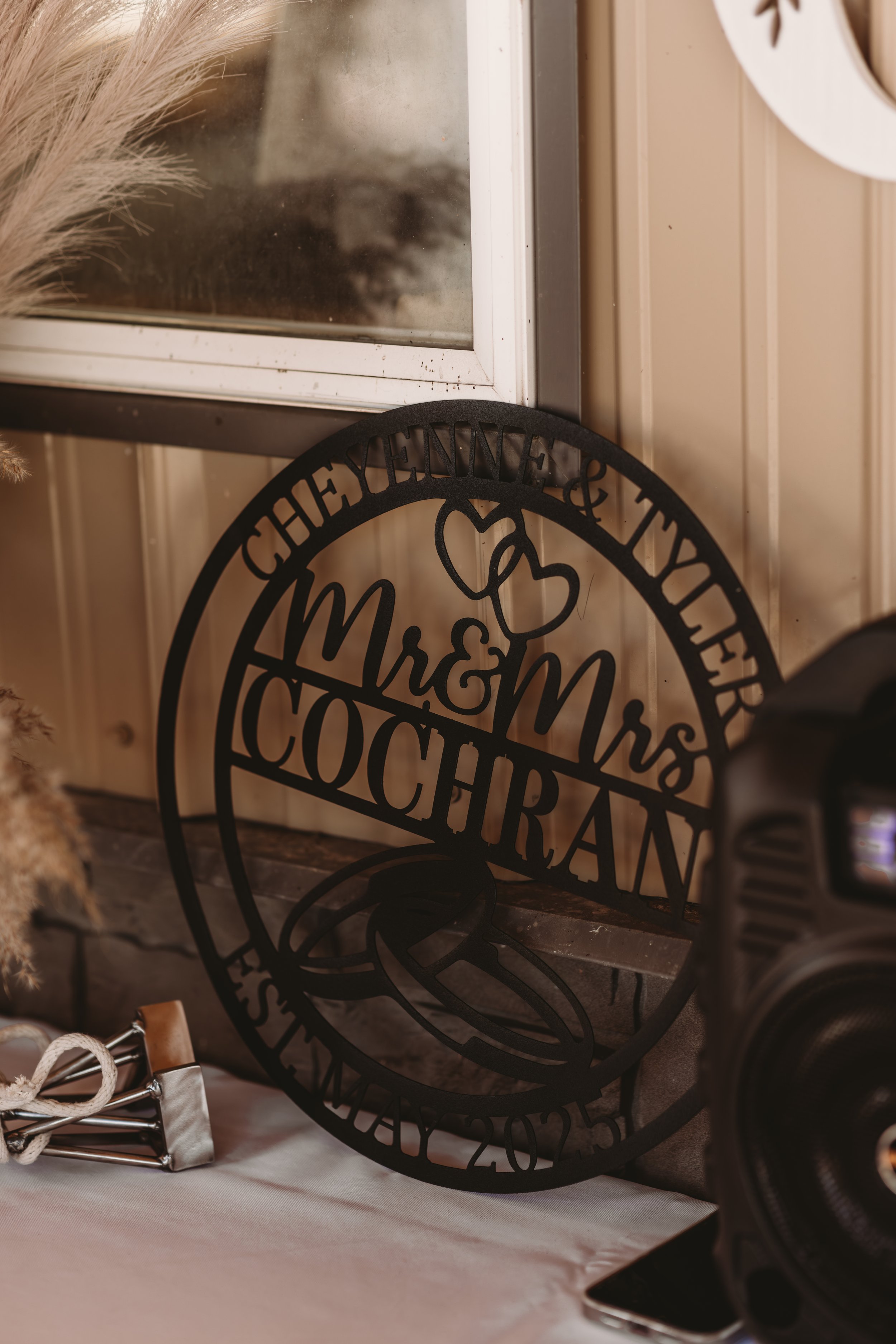 Close-up of a black decorative sign with the words "Mrs. & Mr. Cochran" and hearts, leaning against a window with beige wood paneling. There are festive items on a table below the sign.