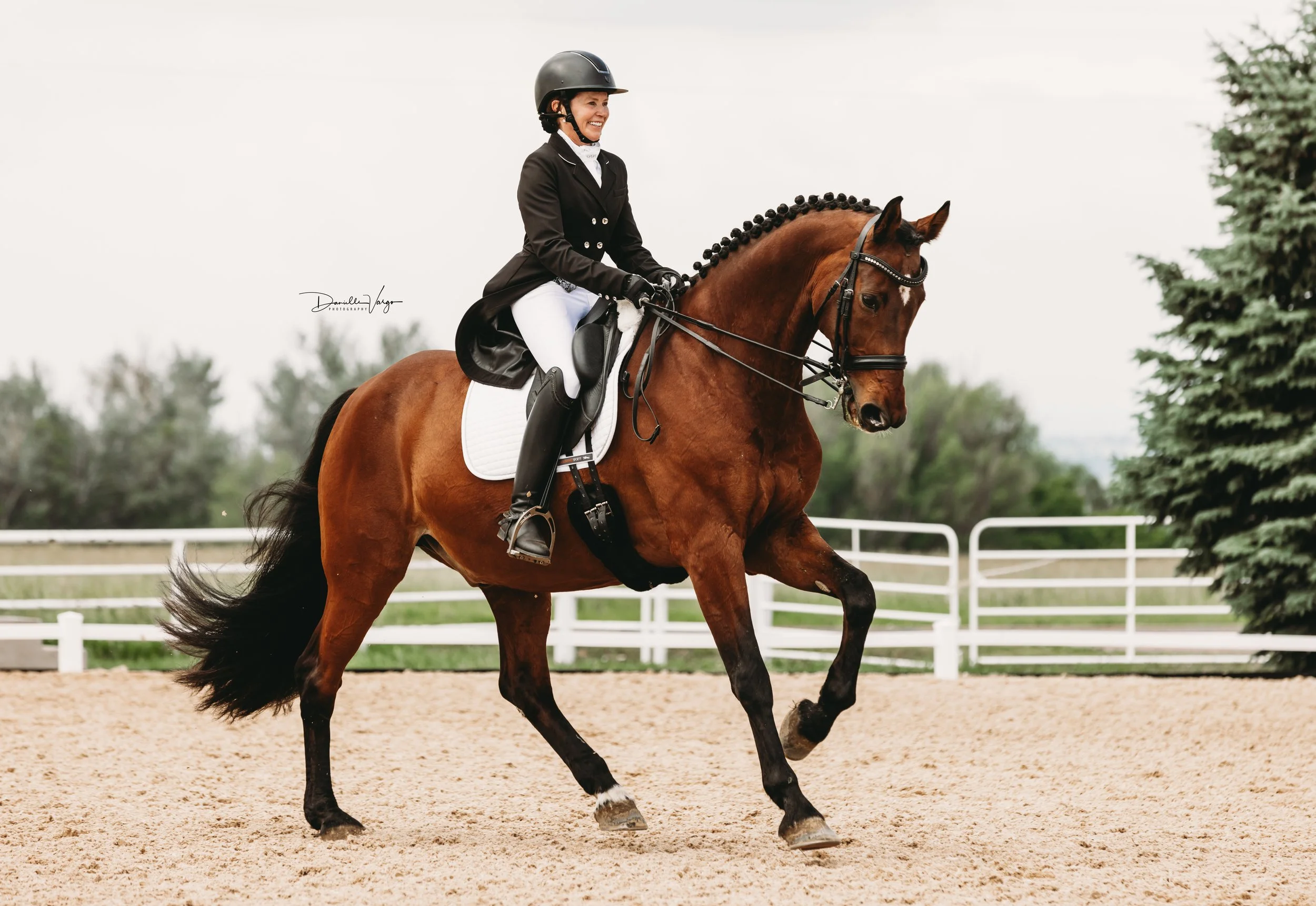 Woman riding a brown horse on a sandy equestrian track, wearing a black helmet and a black riding jacket.