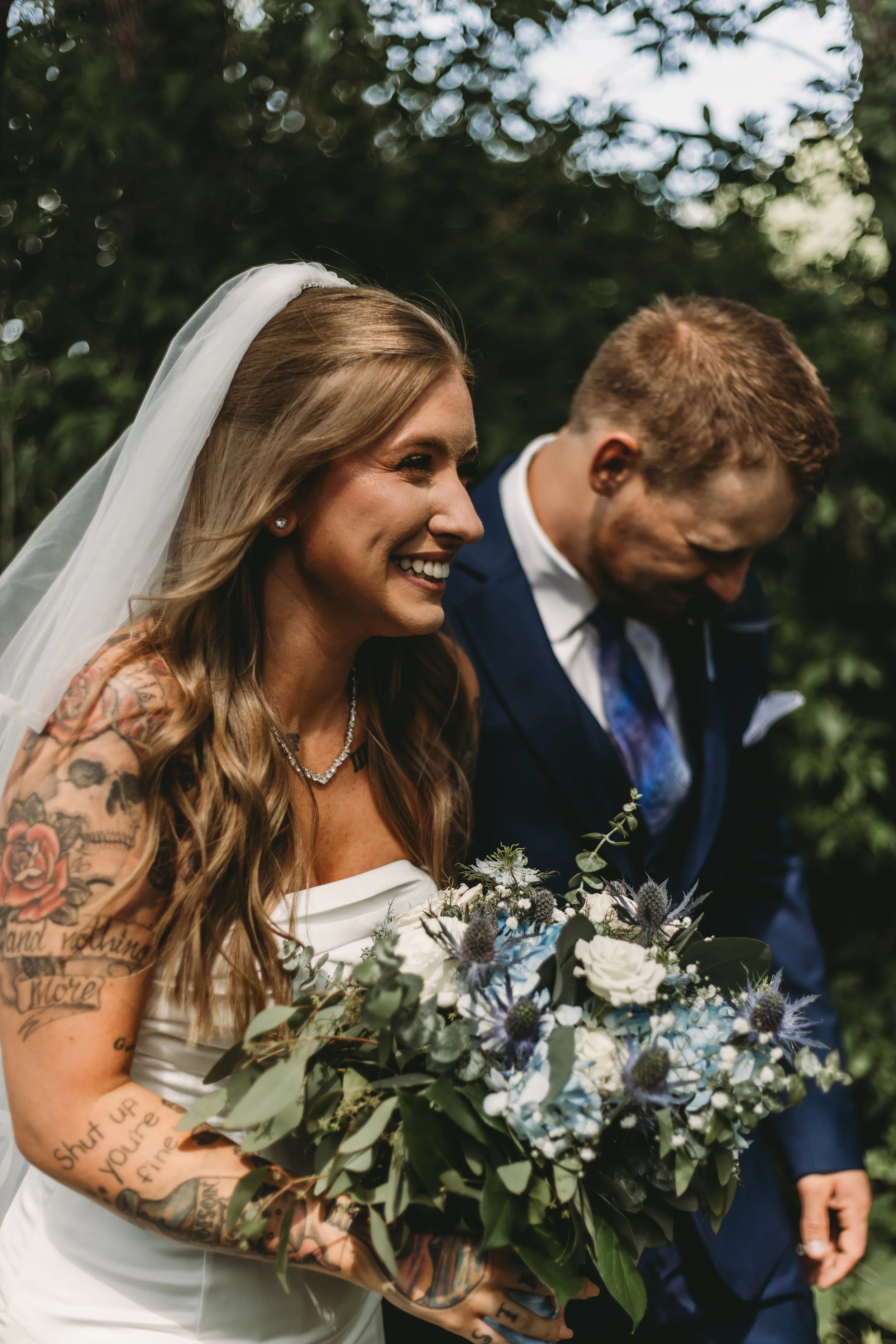A smiling bride with long, wavy hair wearing a white strapless wedding dress and a veil, holding a bouquet of white and blue flowers, standing next to a groom in a blue suit, outdoors with greenery in the background.
