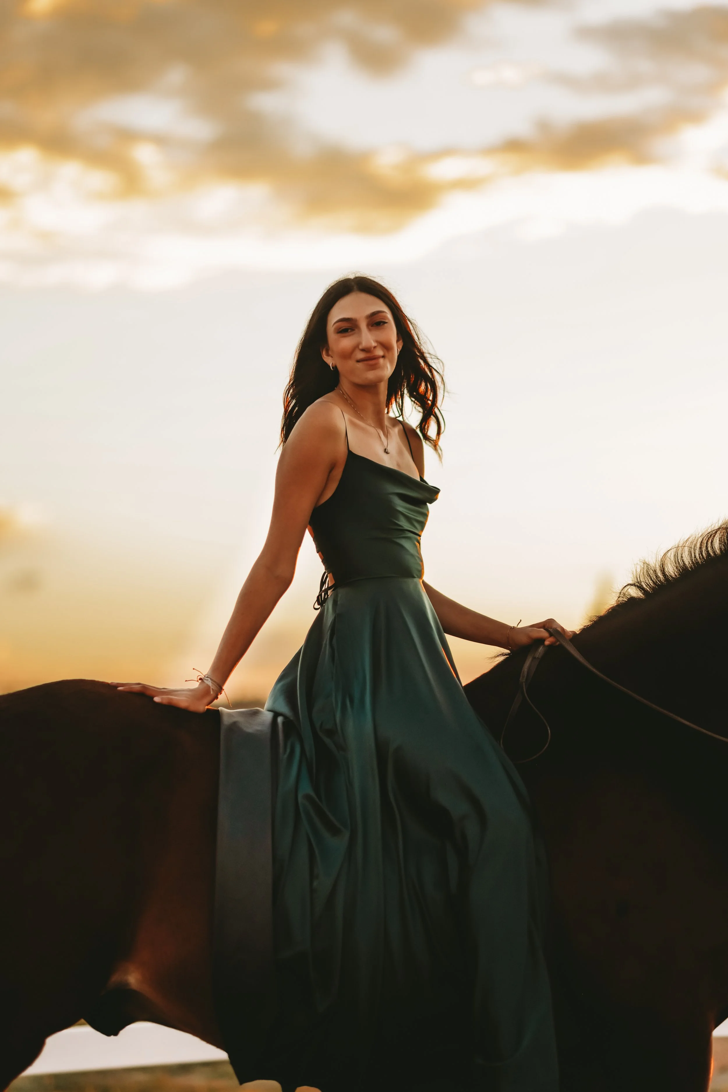 A young woman in a dark green dress riding a horse during sunset, with clouds in the sky.