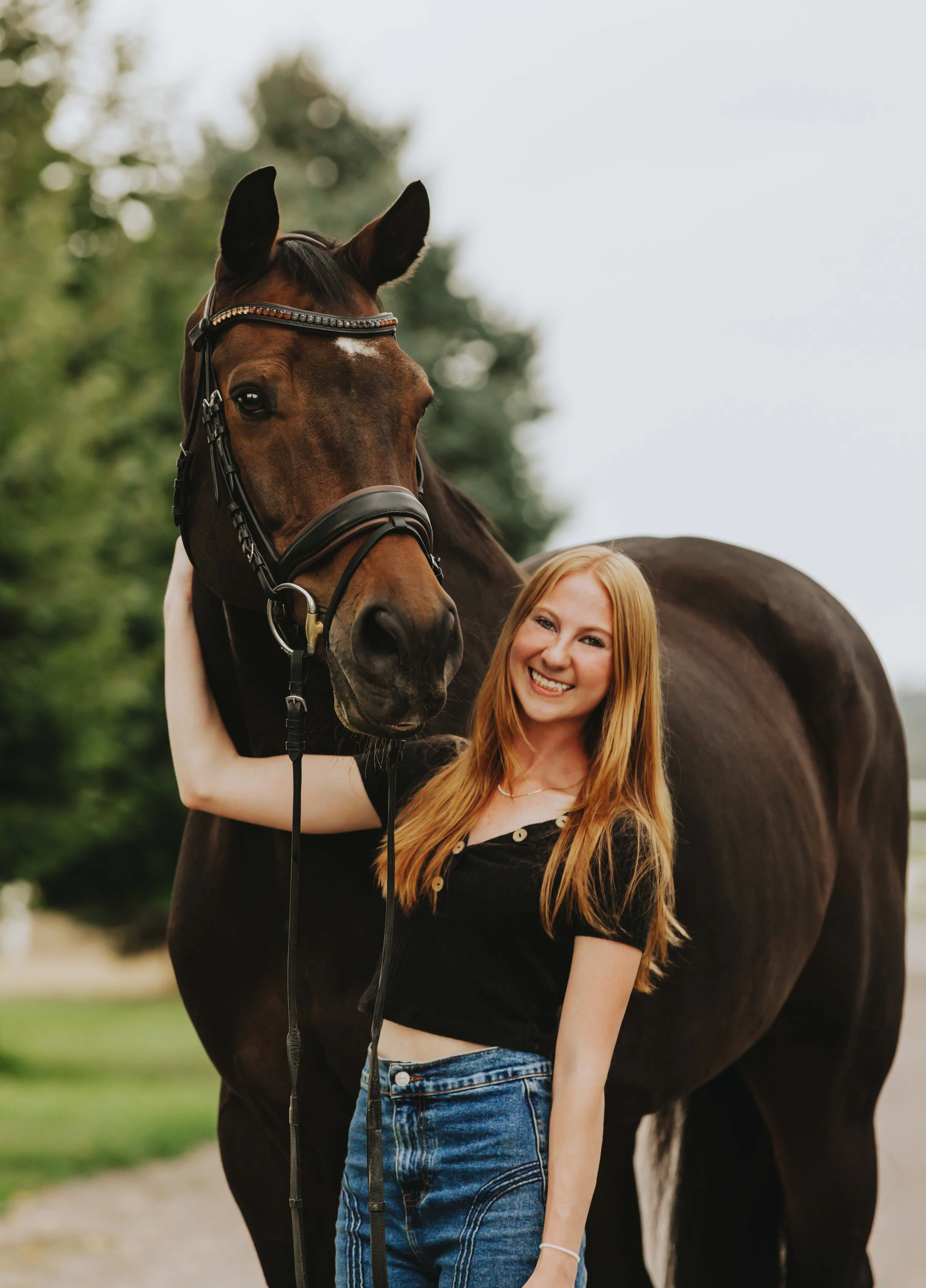 A young woman with long red hair smiling and standing next to a dark brown horse with a bridle, outdoors with blurred greenery in the background.