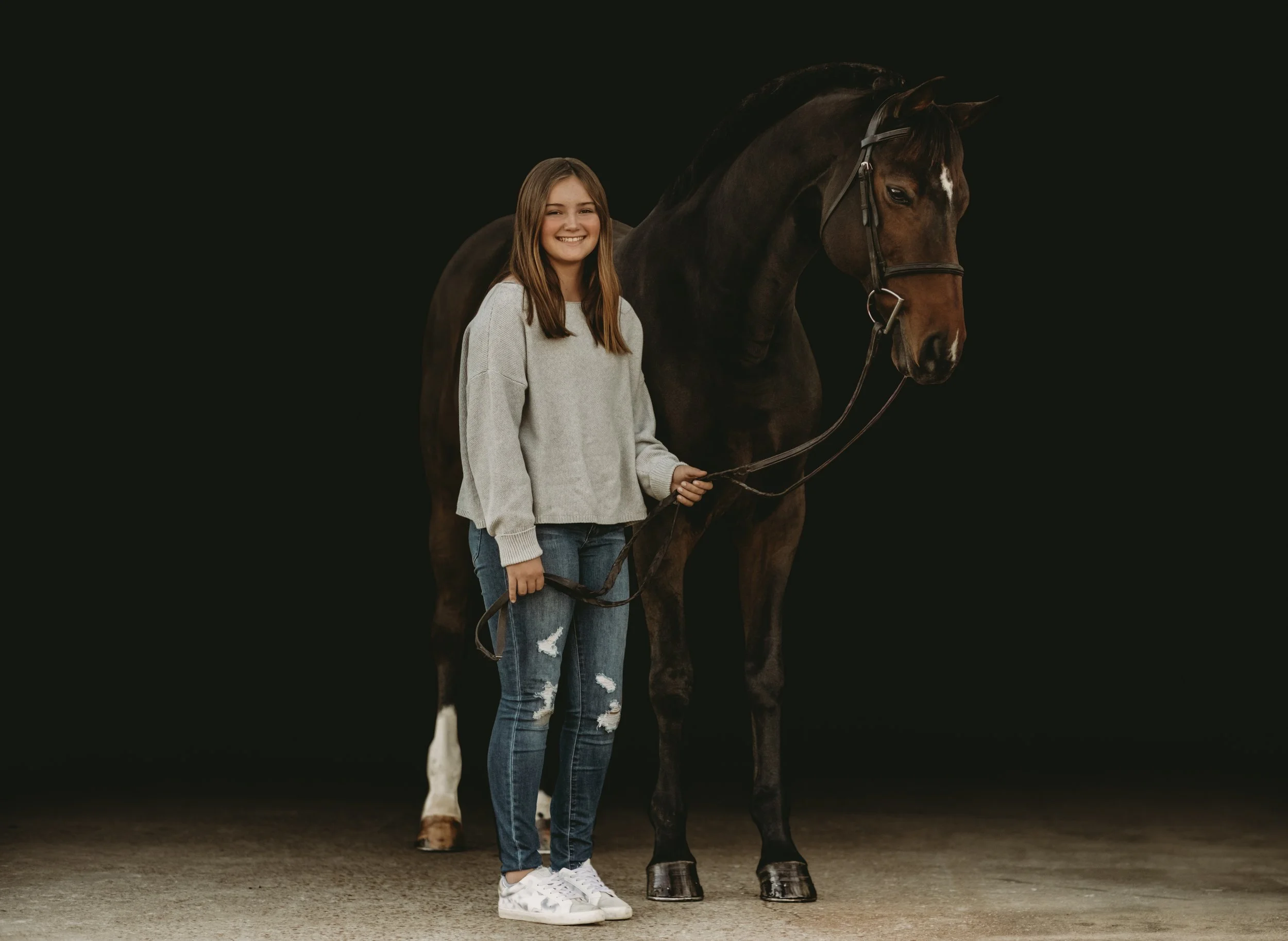 A girl with long brown hair, wearing a light gray sweater and ripped jeans, holding the reins of a dark brown horse with a white star on its forehead, against a dark background.