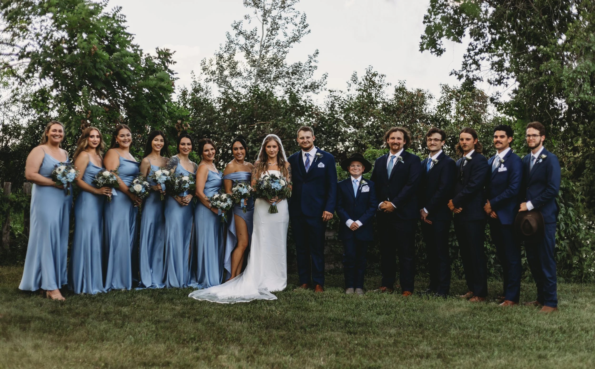 A large wedding party standing outdoors on grass with trees in the background, including bridesmaids in light blue dresses holding bouquets, a bride in a white wedding dress with a veil, and groomsmen in navy suits with boutonnieres, along with a you