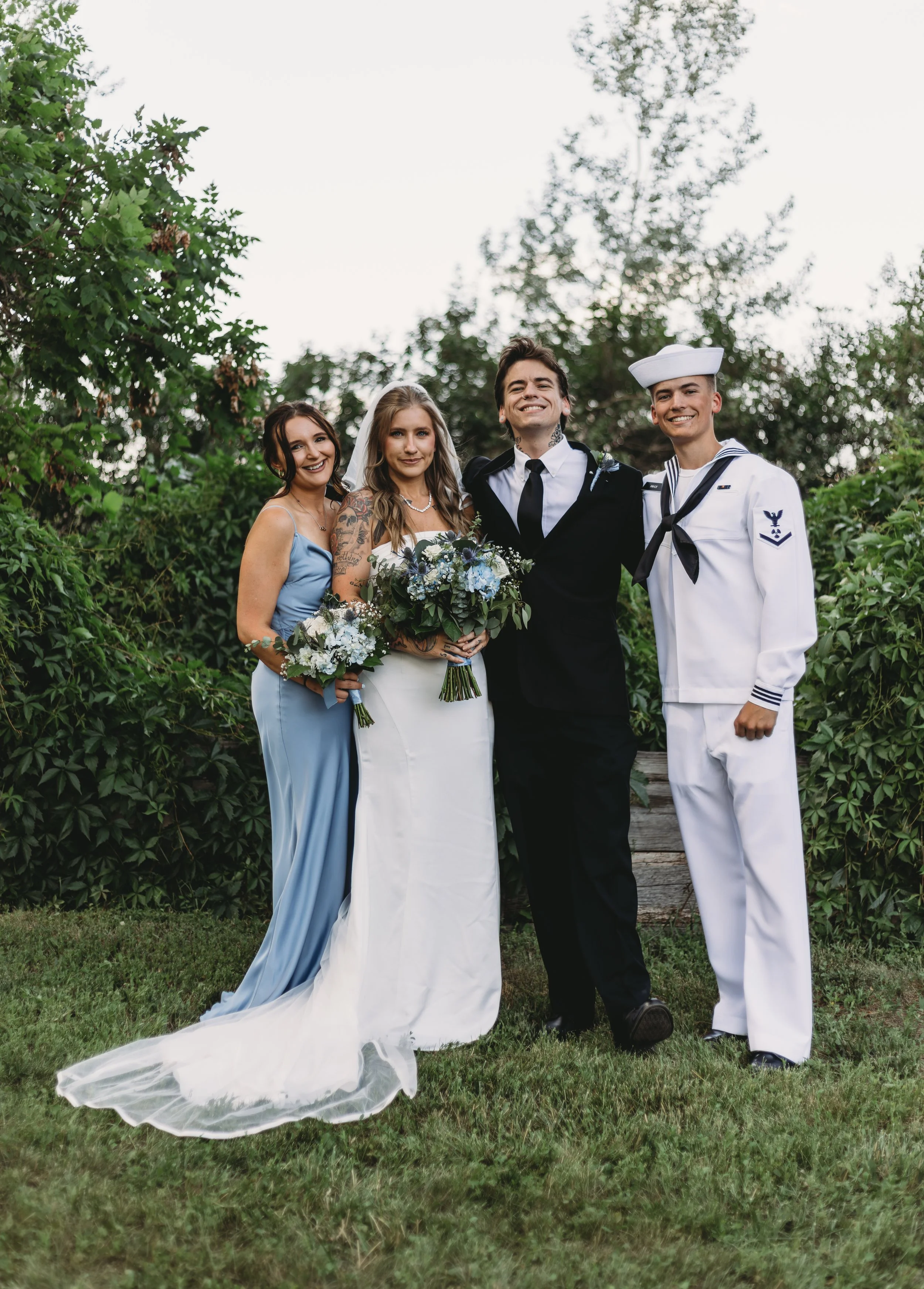 Four people standing outdoors among trees and greenery, dressed in wedding and military uniforms, smiling and posing for the photo.