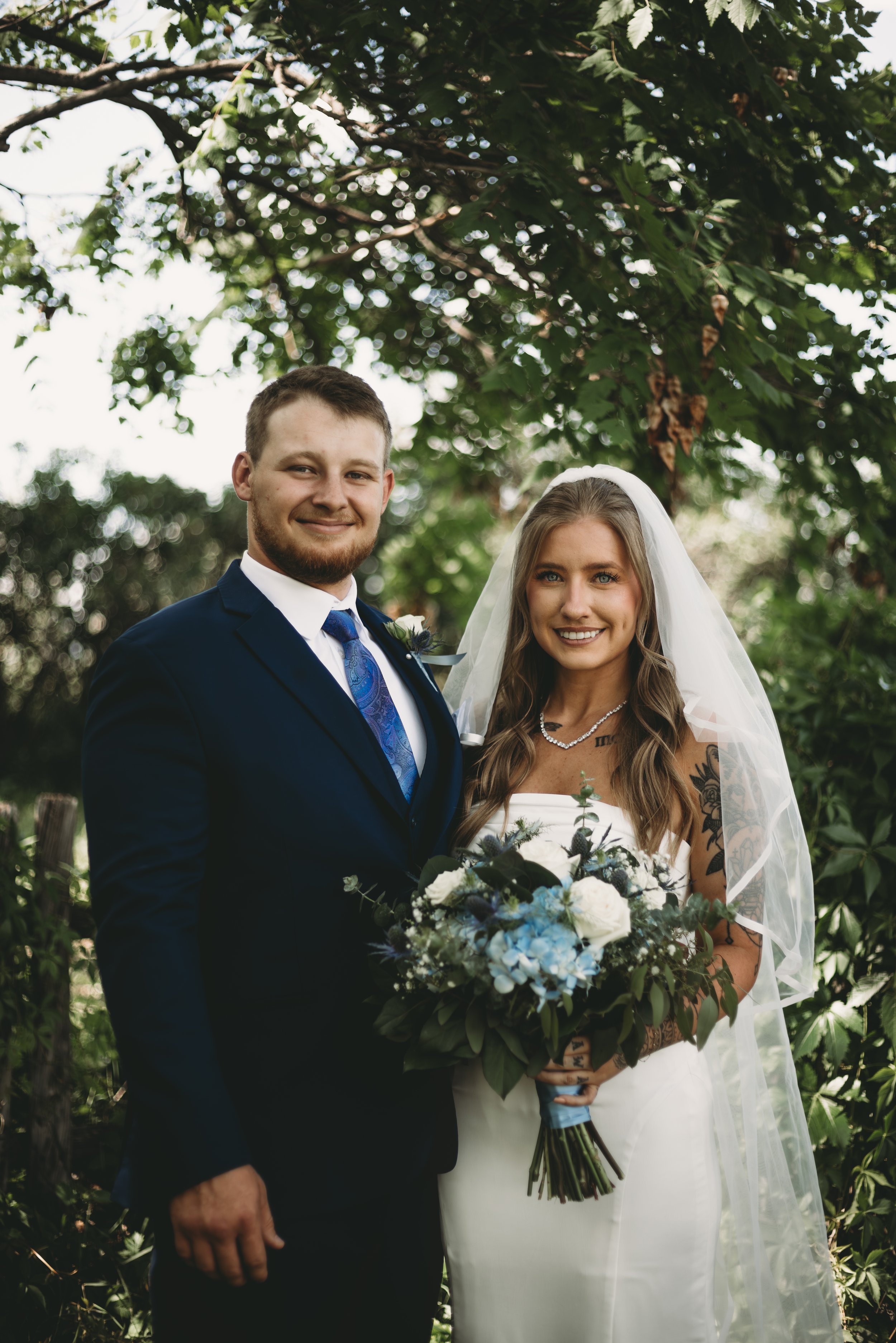 A happy couple dressed in wedding attire standing outdoors in front of a leafy green background. The man is wearing a navy blue suit with a white shirt and blue patterned tie, and the woman is wearing a strapless white wedding dress with a veil, hold