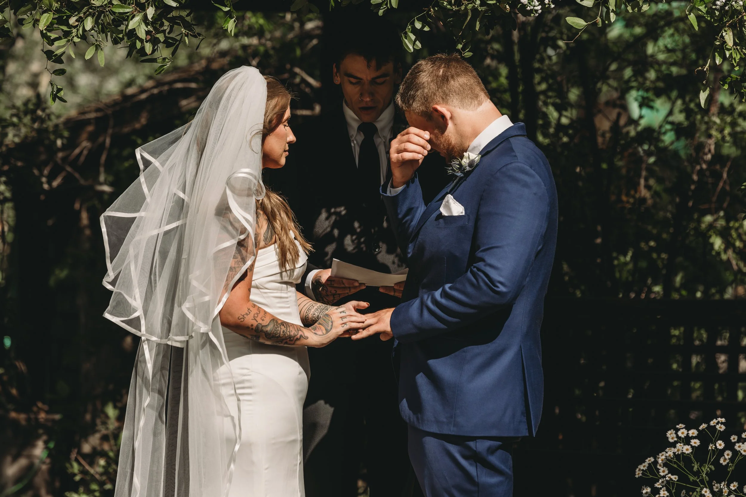 A couple getting married outdoors, holding hands and praying with an officiant during the ceremony. The bride wears a veil and tattooed arms, and the groom is in a blue suit. Green foliage surrounds them.