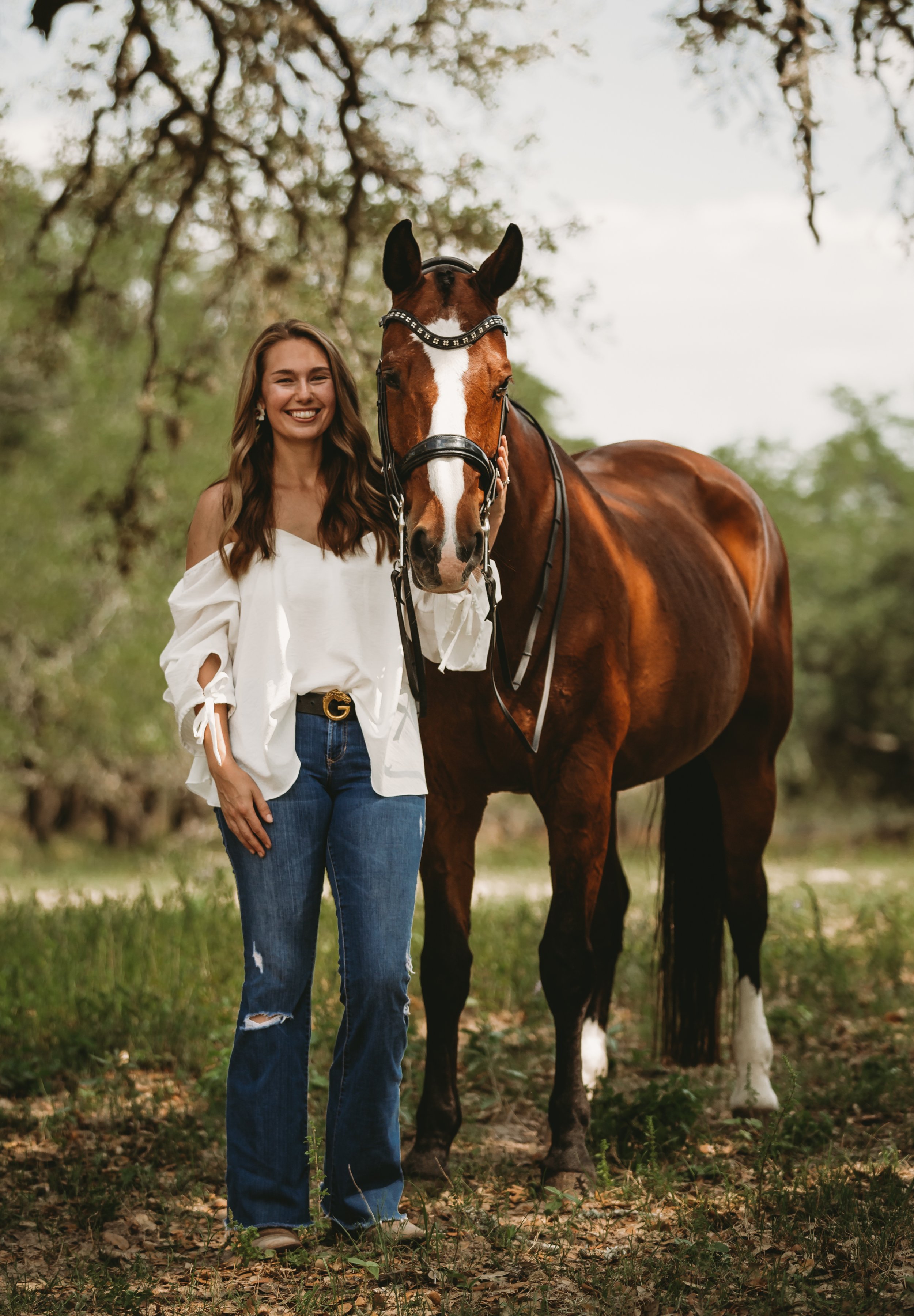 A smiling woman with long brown hair wearing a white off-shoulder blouse and ripped blue jeans standing next to a brown horse with a white stripe on its face, in a wooded outdoor setting.