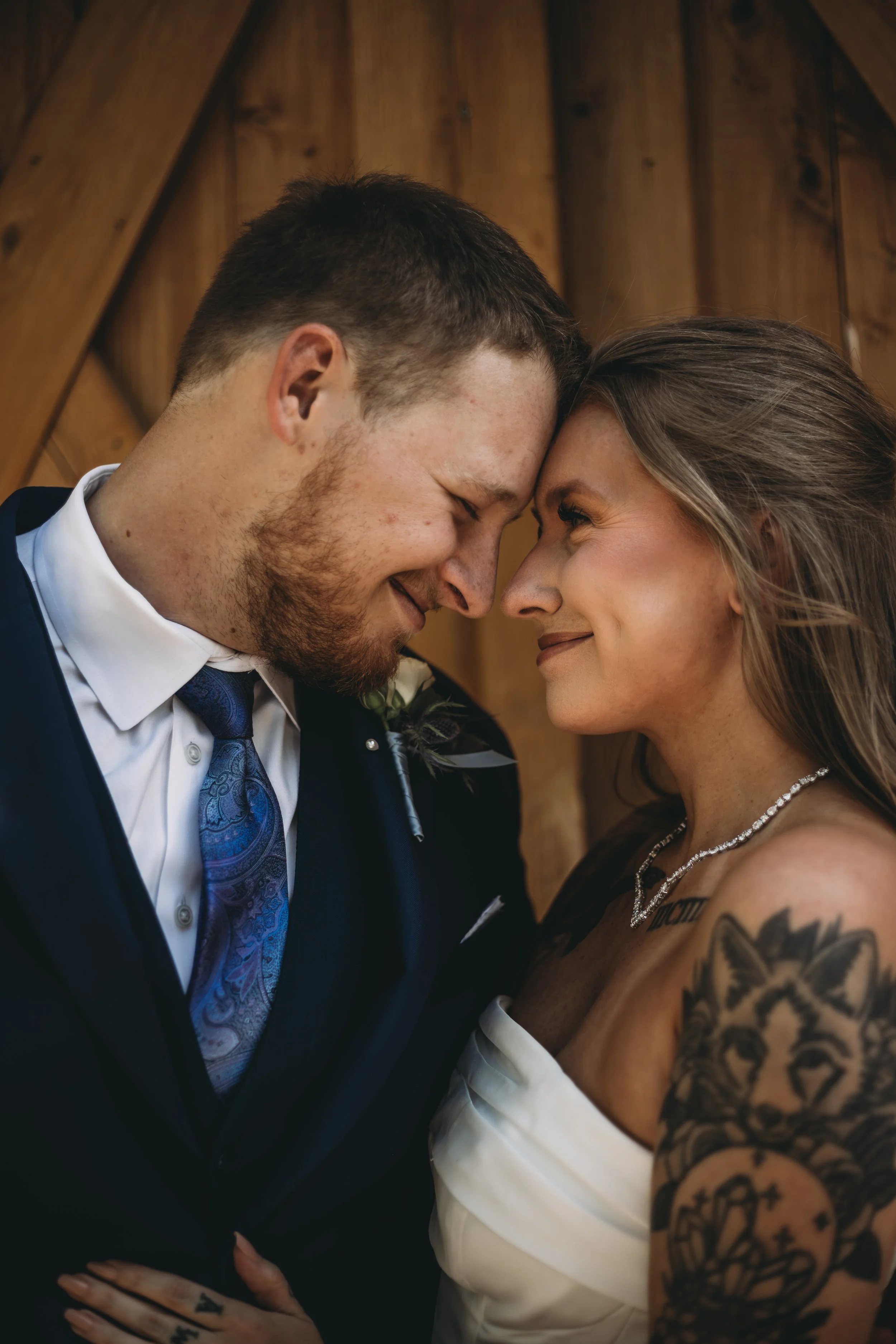 A couple with foreheads touching, eyes closed, smiling, in wedding attire inside a wooden building.