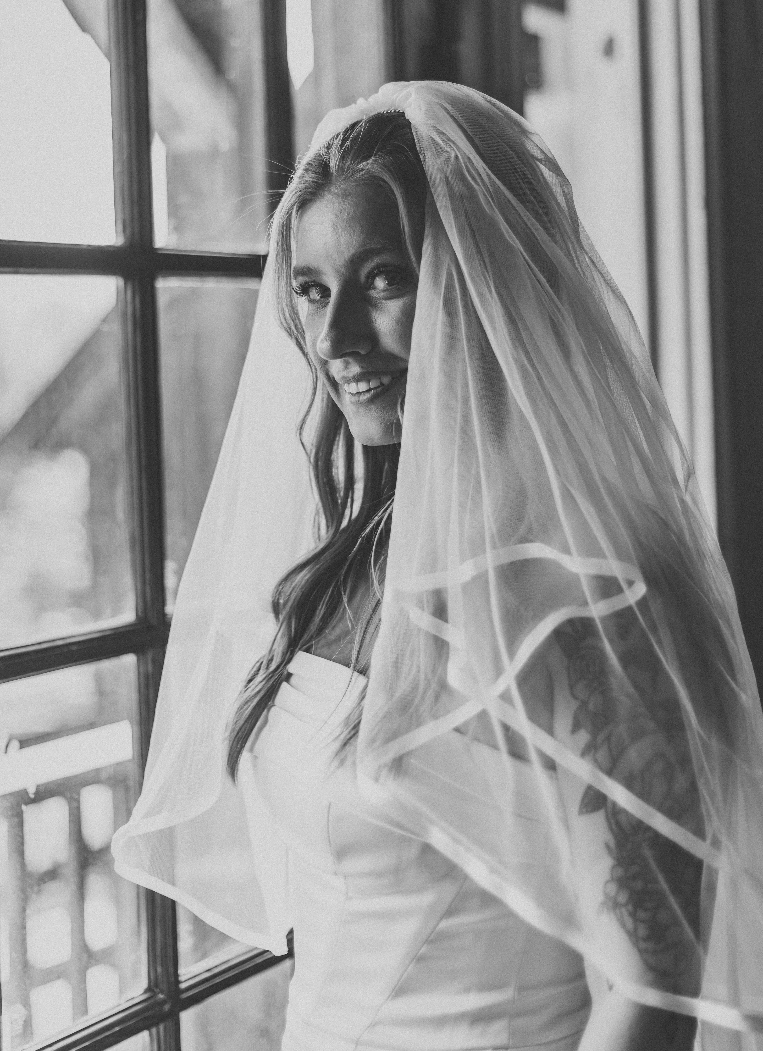 Black and white photograph of a smiling woman with long wavy hair and a sheer veil, standing near window with wooden framing.
