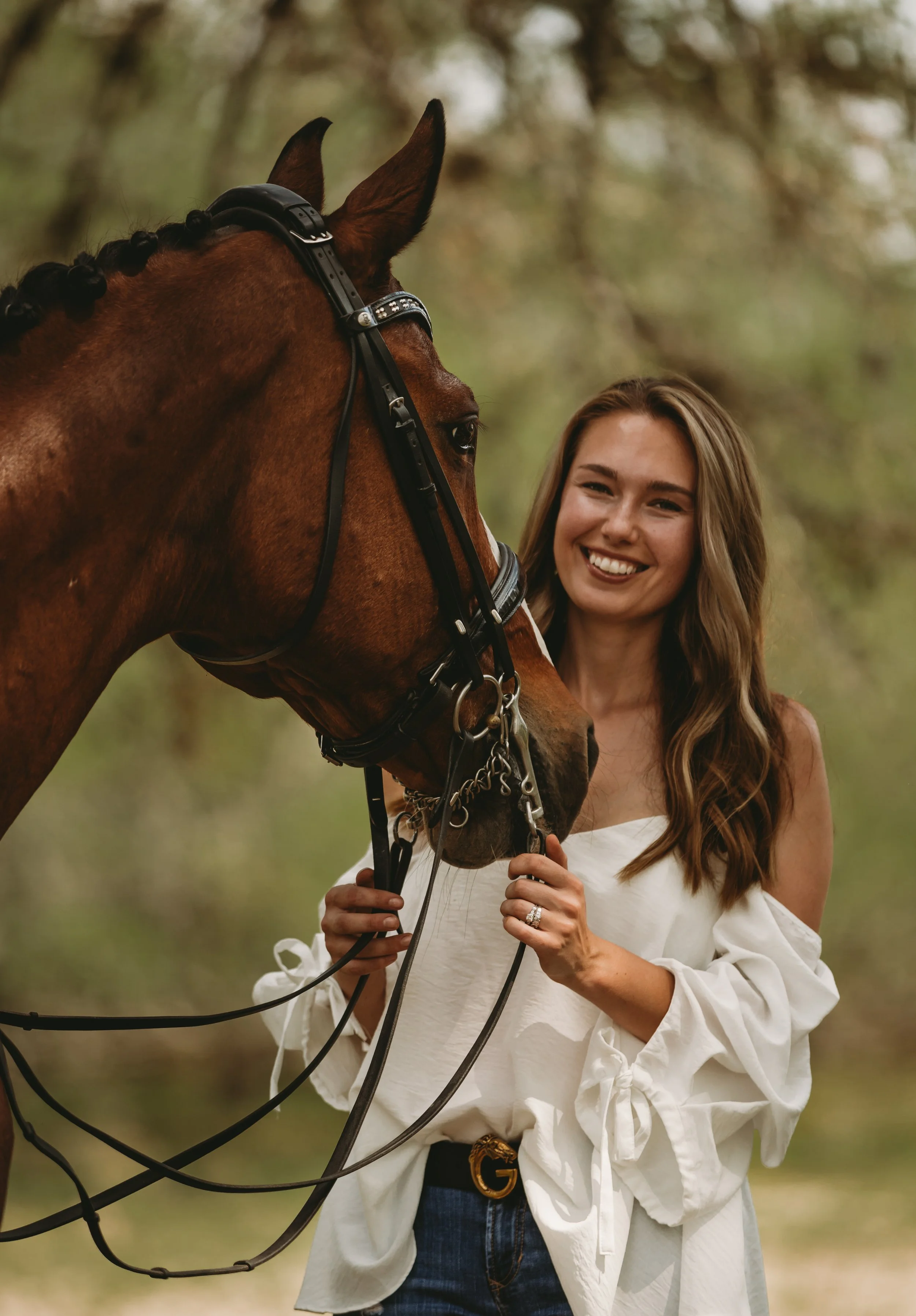 A woman with long, wavy hair smiling while holding a brown horse by the reins outdoors in a wooded area.