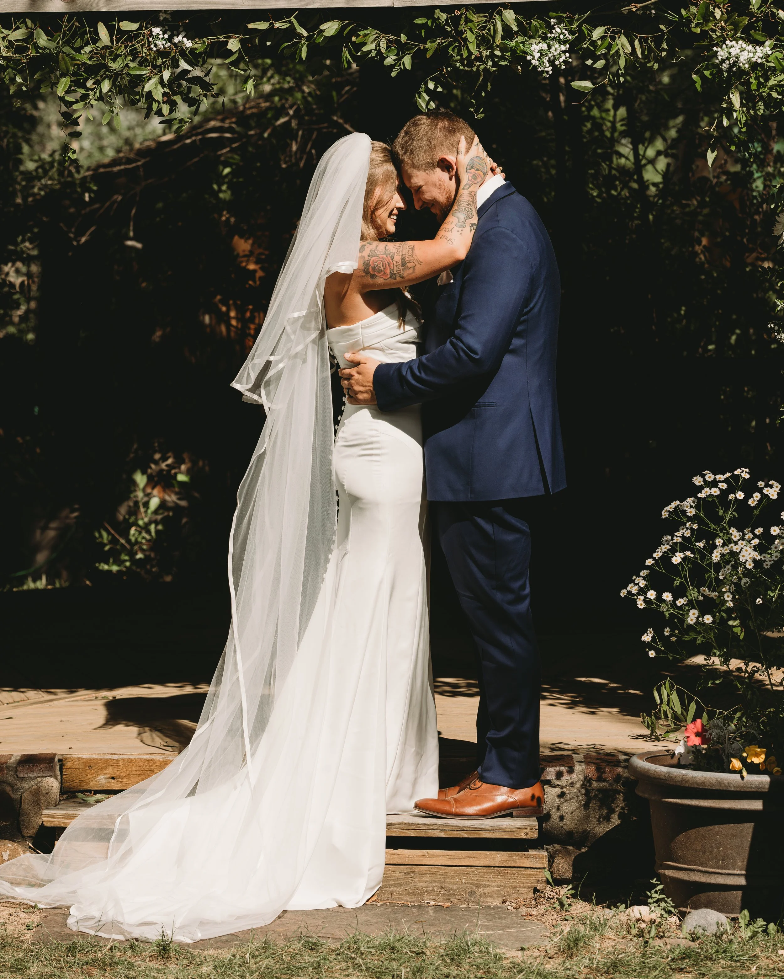 A bride and groom are standing closely together on a wooden platform outdoors, with their foreheads touching and eyes closed, sharing an intimate moment during their wedding. The bride is wearing a white wedding dress and a veil, and the groom is dre