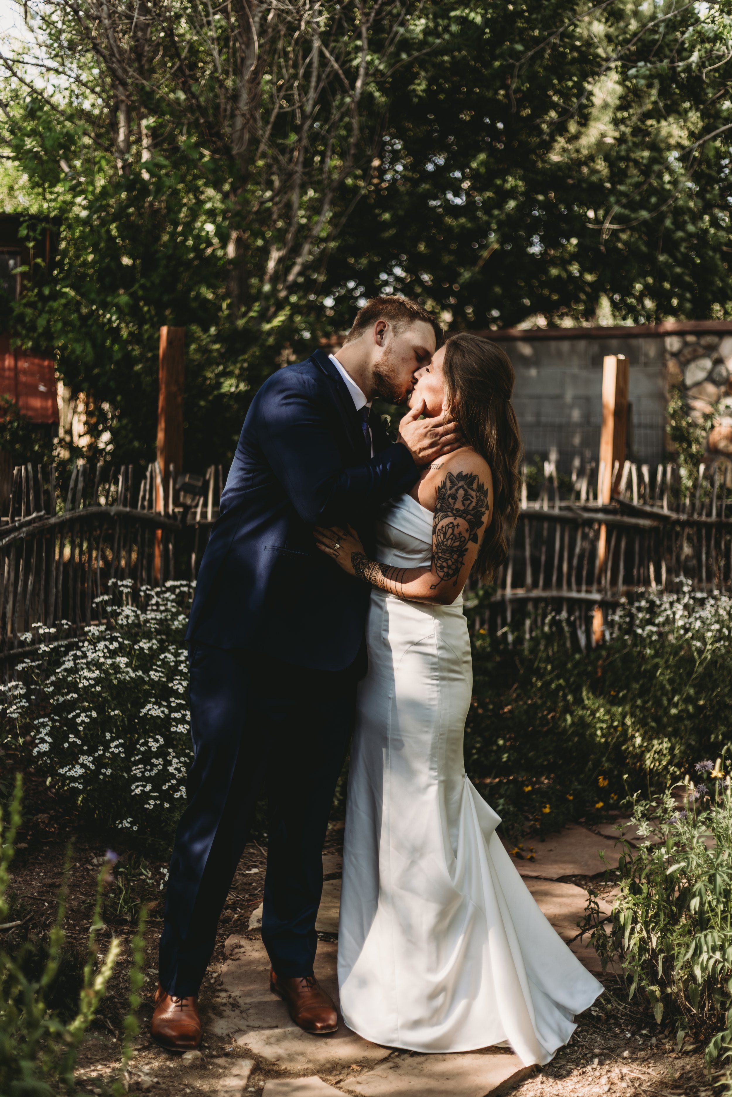 A couple dressed in wedding attire sharing a kiss outdoors surrounded by greenery.