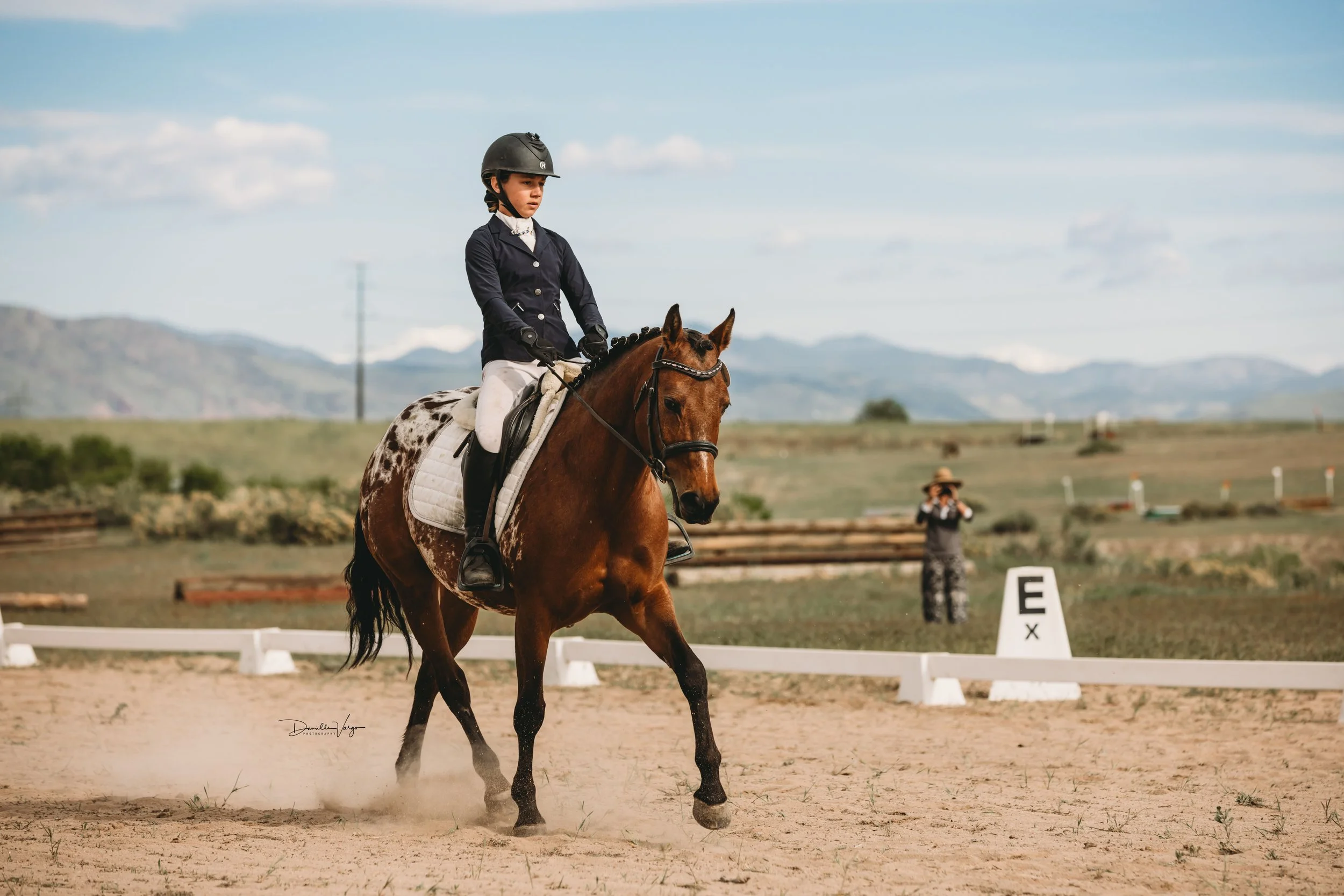 A young female equestrian riding a brown and white pinto horse during a competition in an outdoor arena, with spectators and mountain scenery in the background.