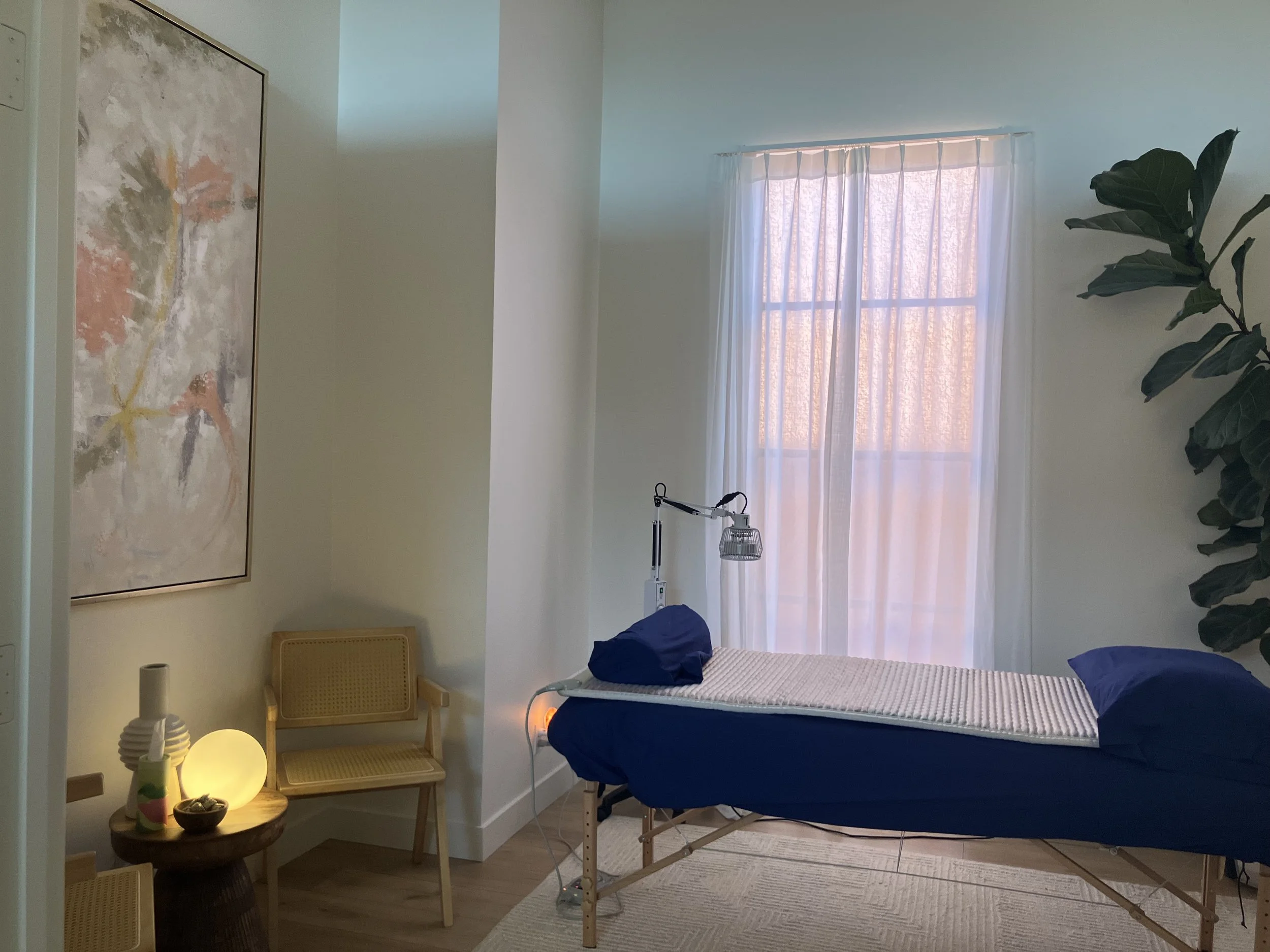 acupuncture treatment room with a massage table, crystal infrared mat on massage table, TDP lamp in back ground, abstract painting on wall, sitting area with wicker chair