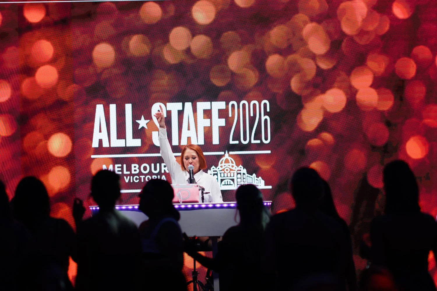 A woman with red hair speaking at a podium during the All-Star TAF 2026 event in Melbourne, Victoria, with a large screen behind her displaying event details and a silhouette of the cityscape.