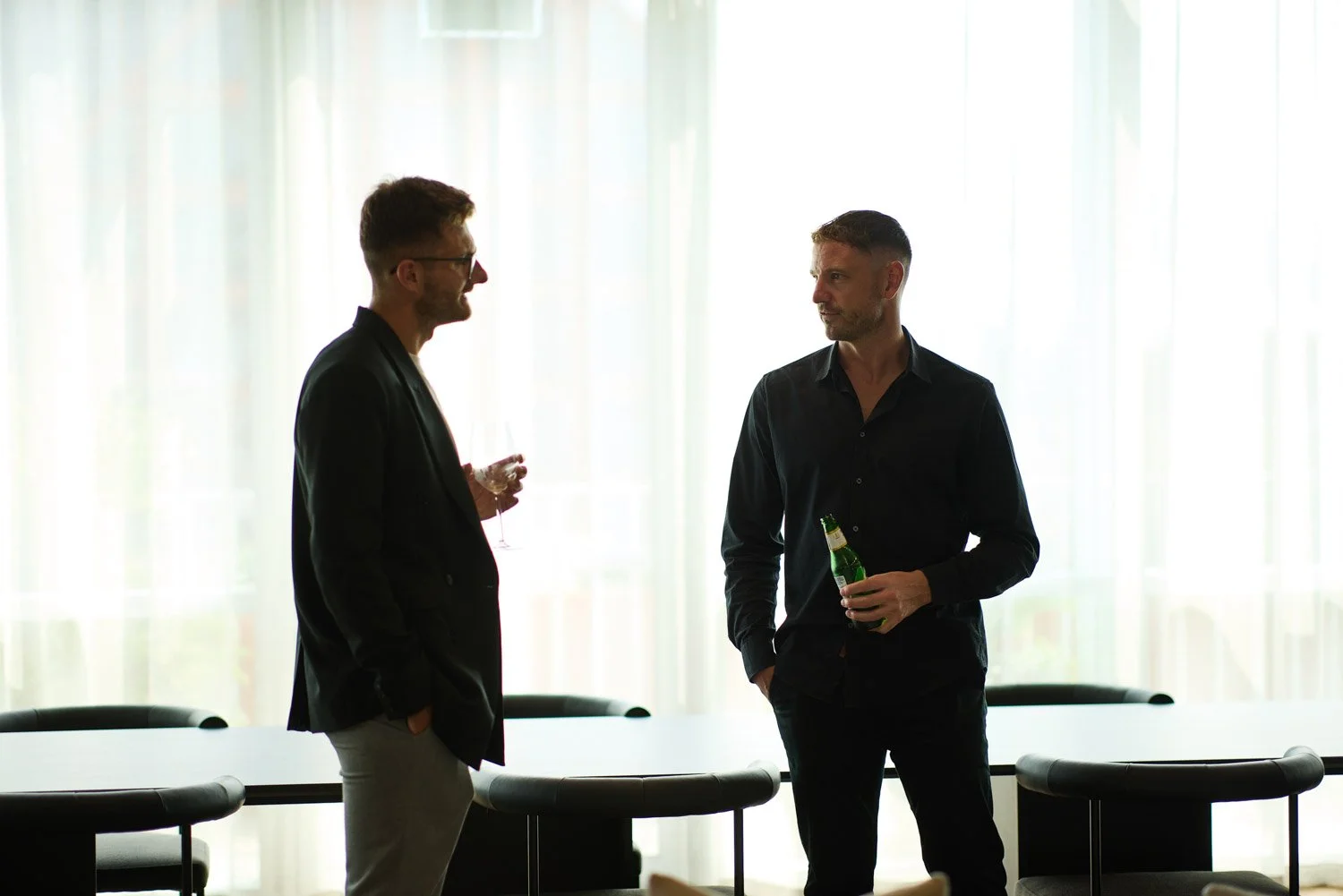 Two men conversation in a room with bright curtains, one holding a glass of wine, and the other holding a beer bottle.