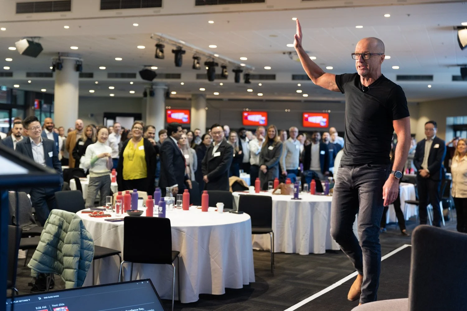 A man in casual black attire and glasses gestures with his right hand on stage in front of a large audience in a conference room.