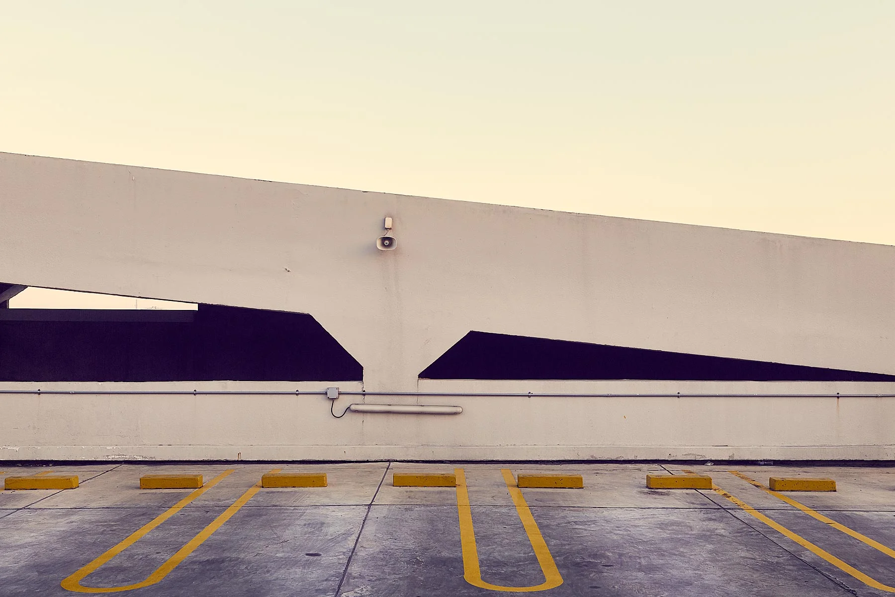 Empty parking lot with yellow painted parking space lines and yellow wheel stops in front of a plain white building with black shaded alcoves, a security camera, and exposed electrical conduit.