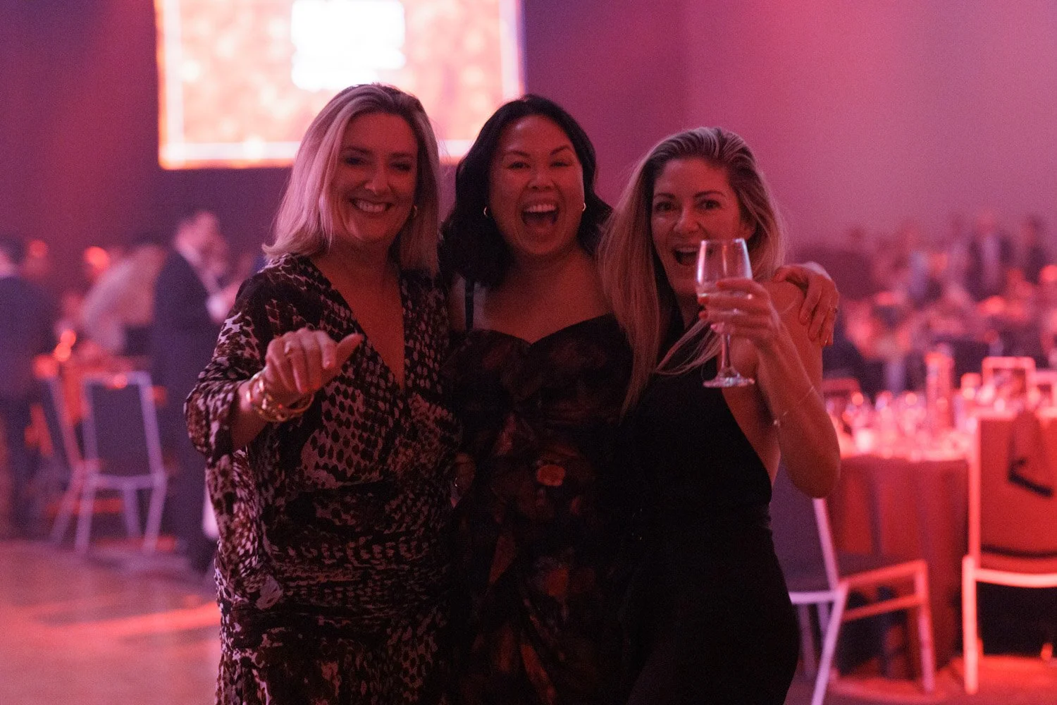 Three women smiling and celebrating at a party, one holding a glass of wine, in a dimly lit event hall with pink and purple lighting.