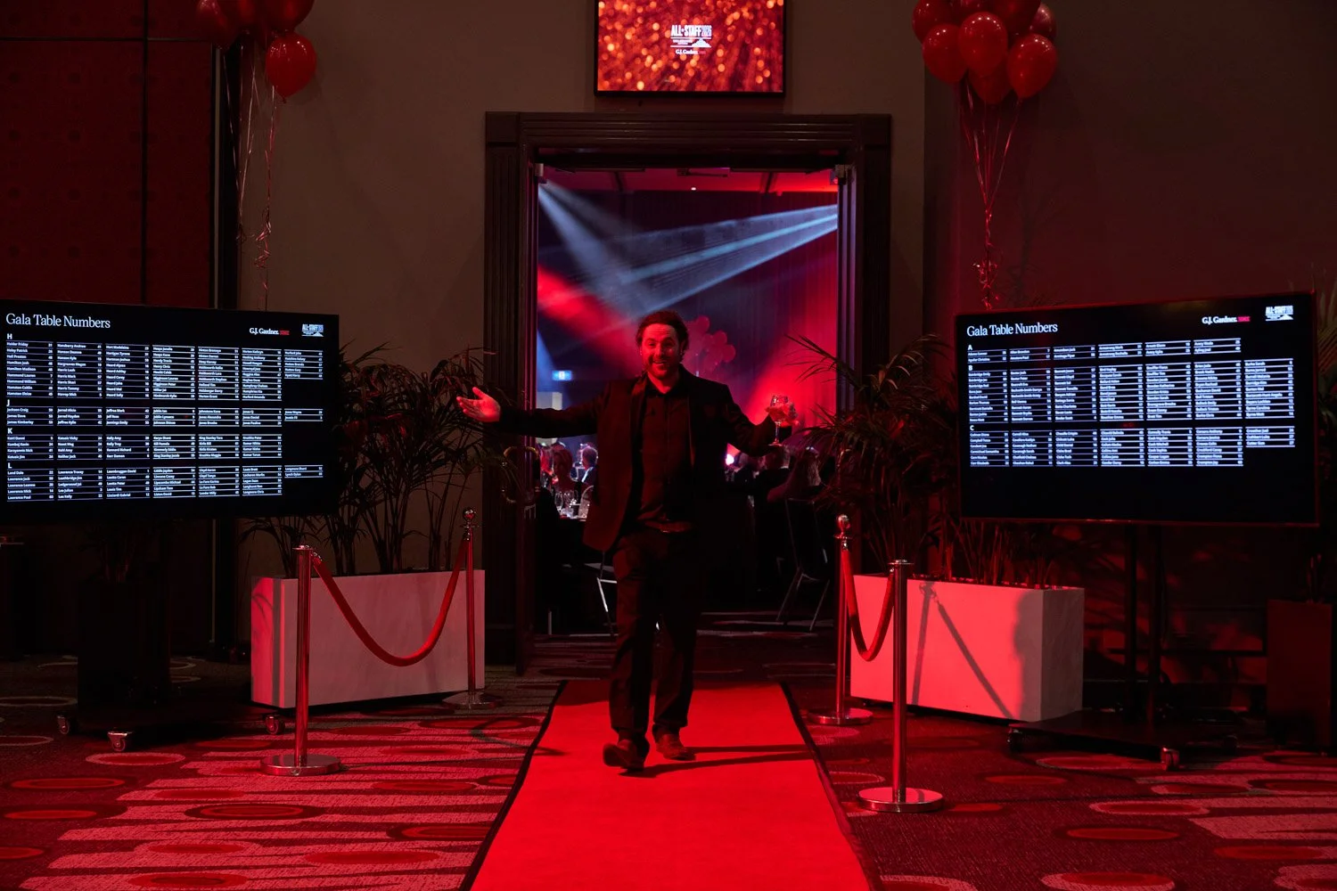 A man in a black suit stands on a red carpet with his arms outstretched, holding a drink in one hand, at an event. Behind him, a doorway leads into a dimly lit room with red lighting, and a large screen displays the text 'All-Star Gala'. Two screens 