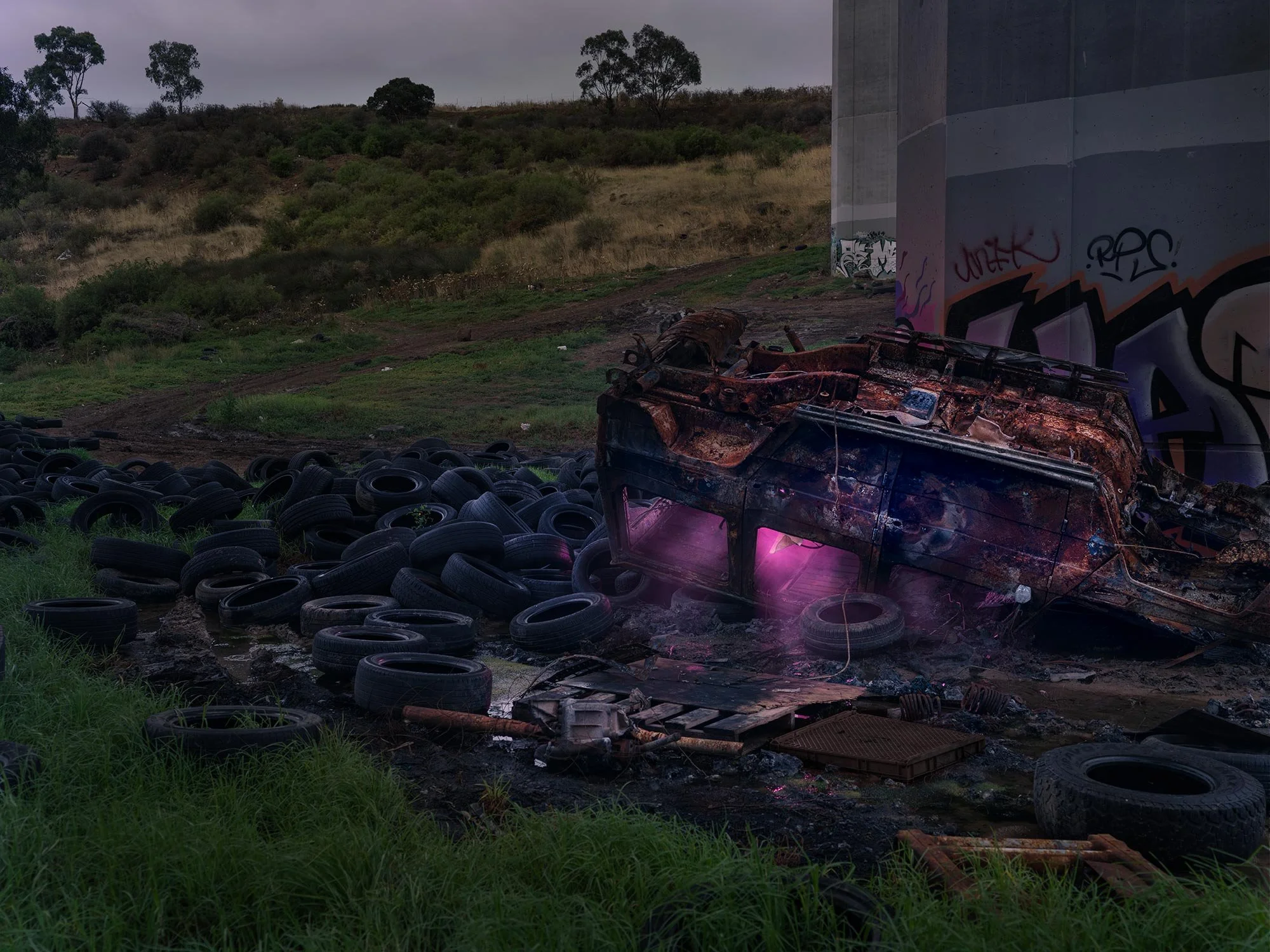 Burned-out vehicle among scattered tires under a bridge with graffiti, surrounded by grassy and hilly terrain.