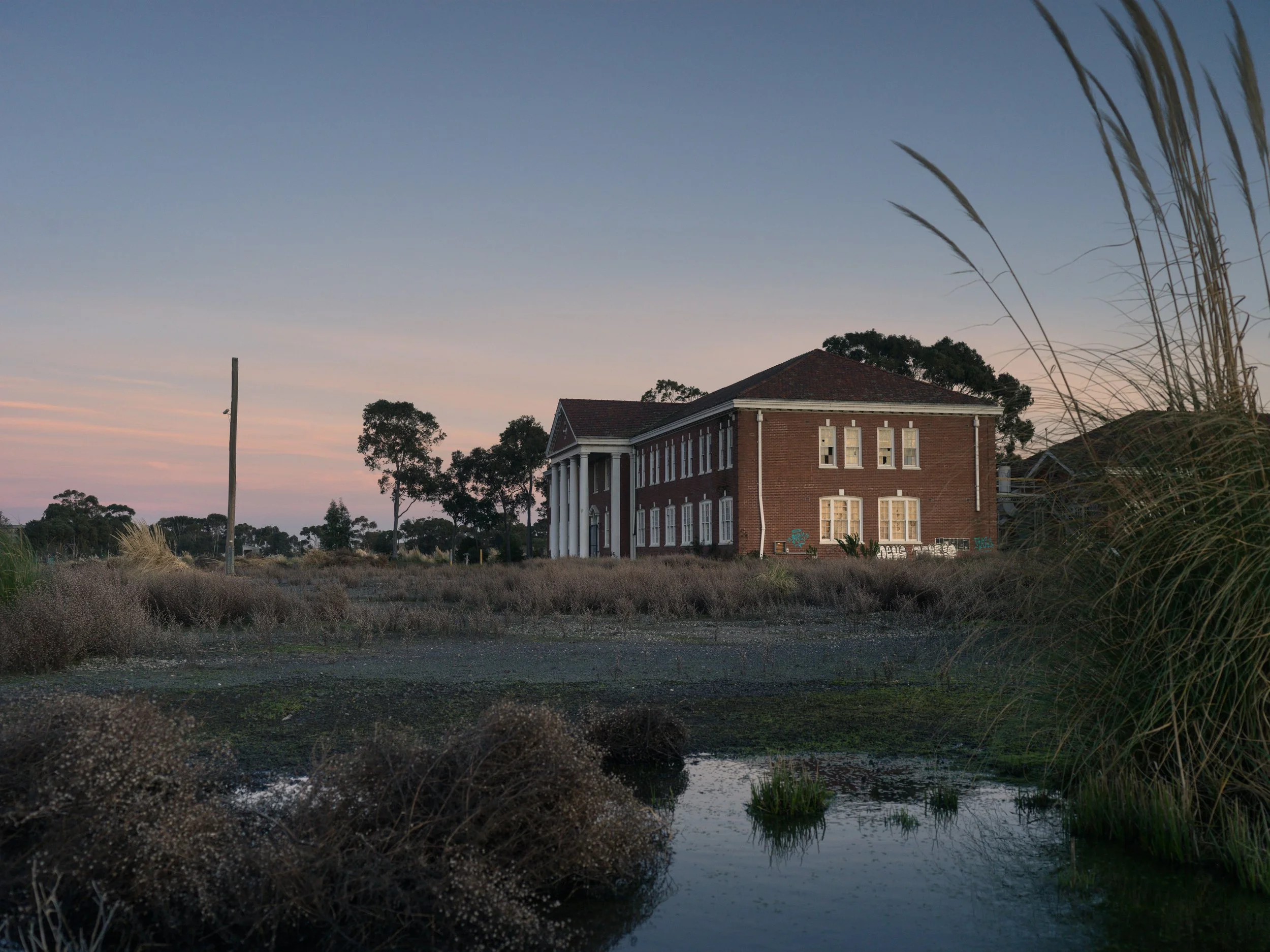 A large brick building with a gabled roof and white columns in the evening, surrounded by grass and trees, with a small water body in the foreground and a clear sky at sunset.