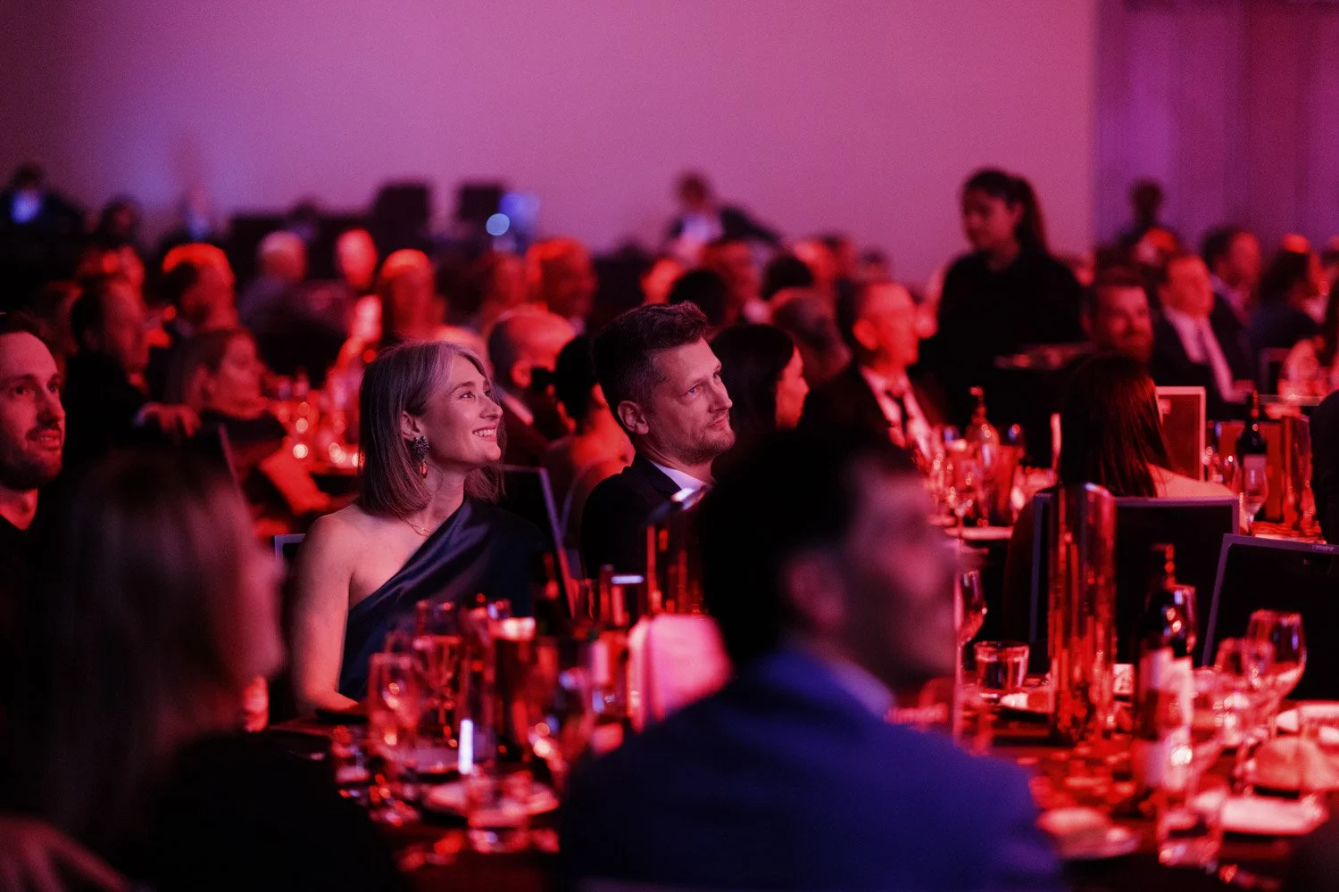 Attendees at a formal event, dressed in elegant clothing, seated at tables with glasses and bottles, under dim pinkish lighting, attentively listening to a speaker or presentation.