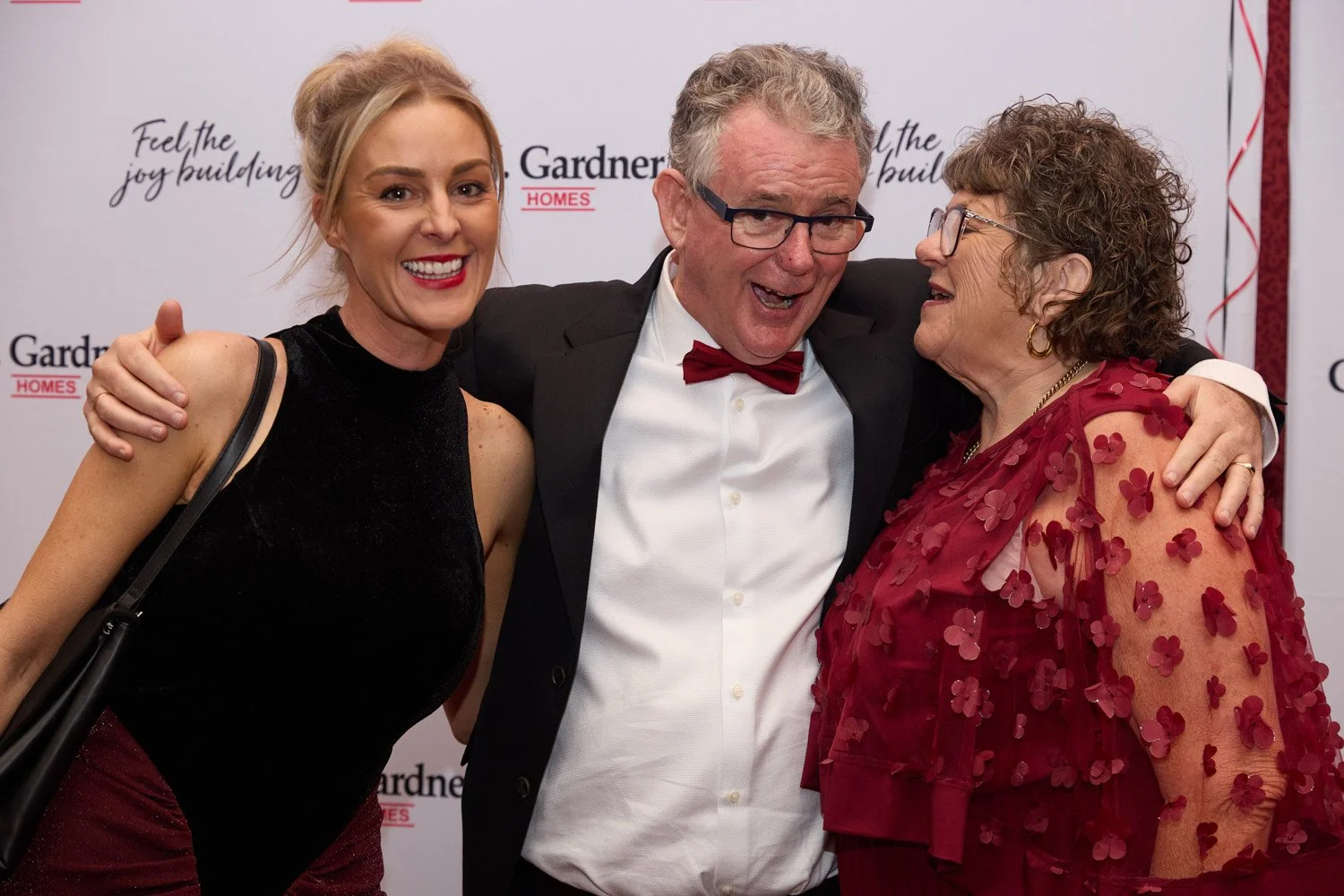 Three people celebrating together at an event, smiling and laughing. The woman on the left has blonde hair in an updo and is wearing a black dress. The man in the middle has curly gray hair, glasses, a black tuxedo with a red bow tie. The woman on th