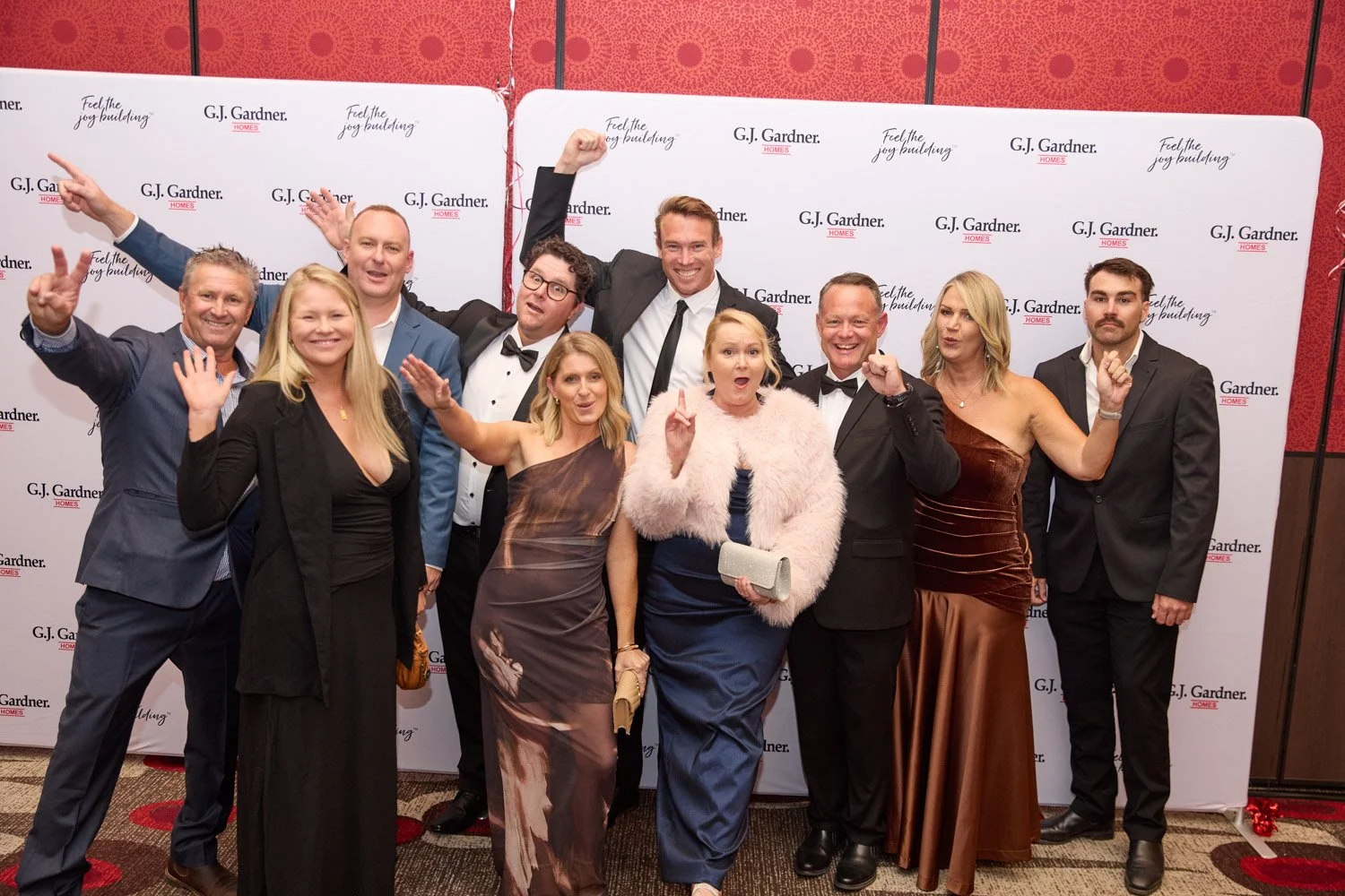 Group of eleven people in formal attire posing on a red carpet in front of a white backdrop with G.J. Gardner Homes logo. Some are smiling, with a few making celebratory gestures.