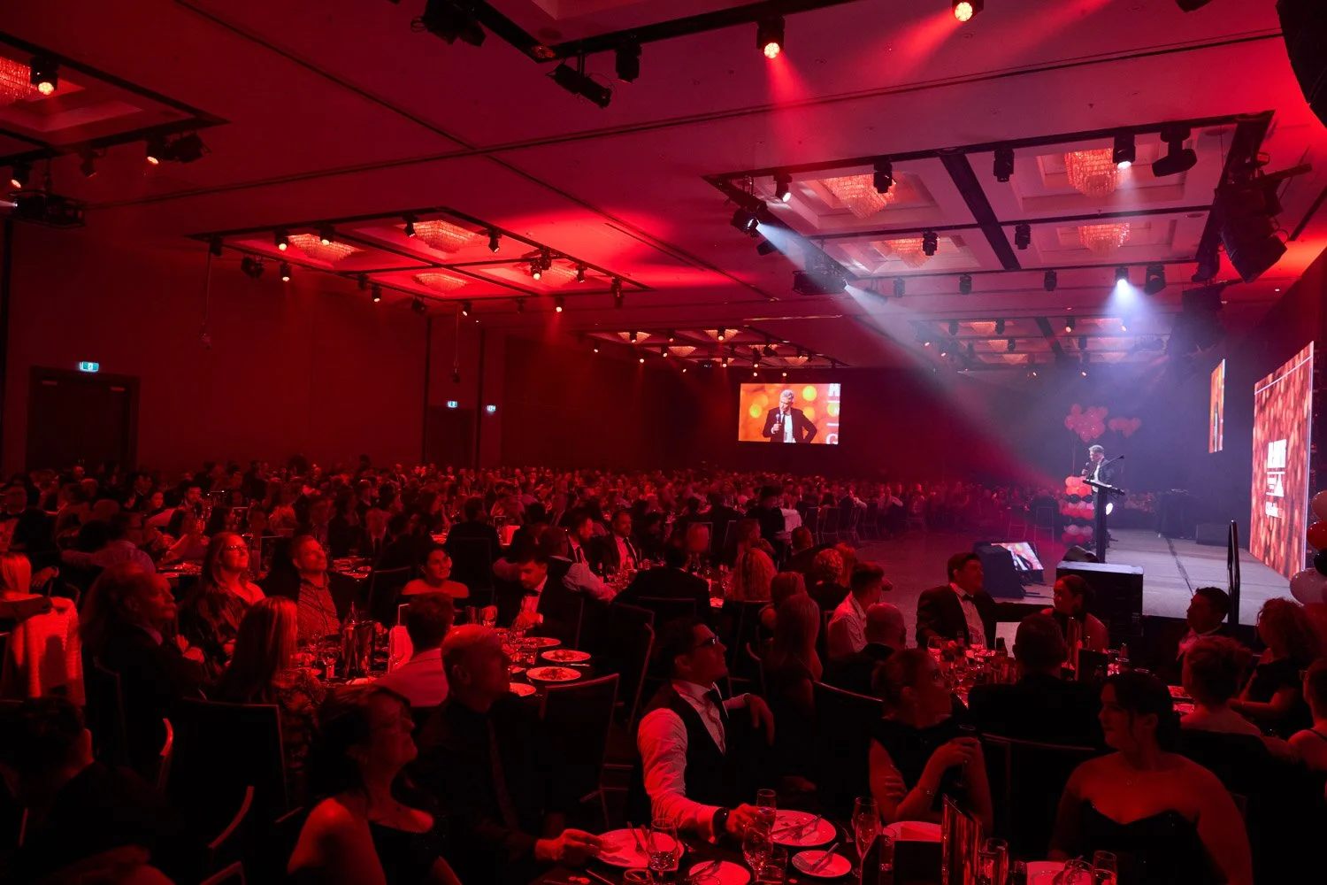 A large banquet hall filled with guests seated at tables, attending a formal event with a stage, speaker, and large screens in the background, illuminated with red and white lights.