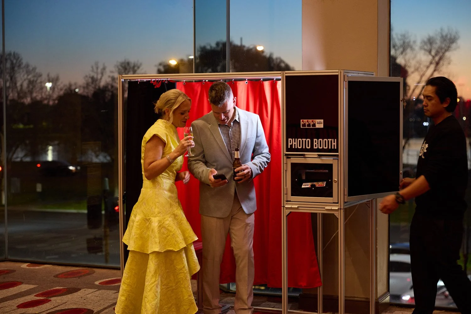 A woman in a yellow dress and a man in a gray suit inside a photo booth with red curtains, while another man in black stands outside.