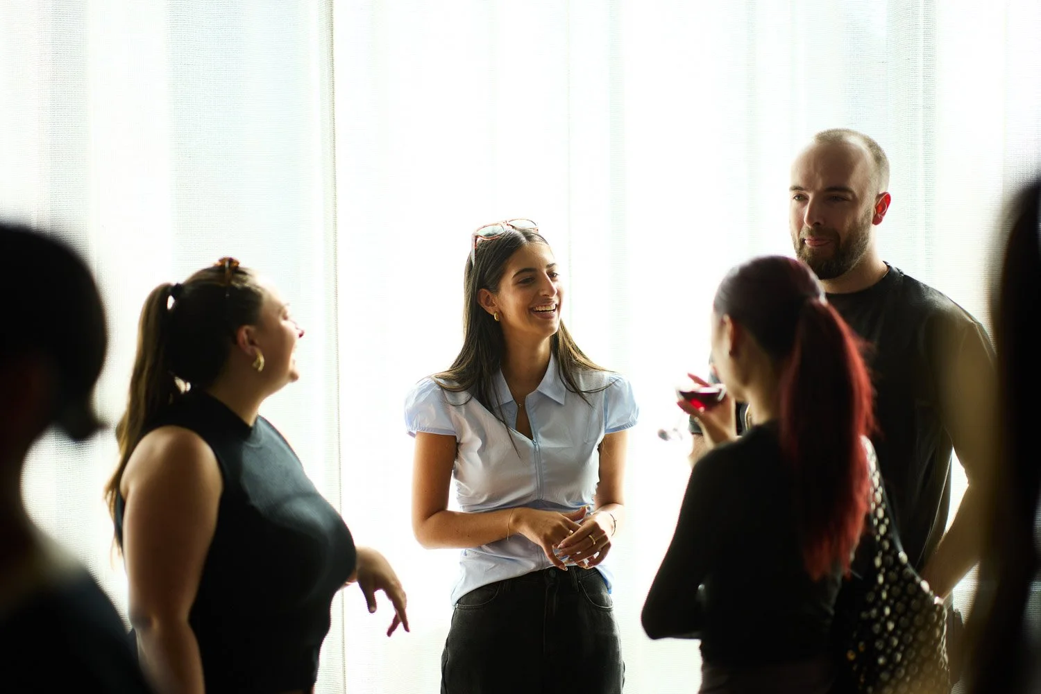 Group of five people talking and laughing together in front of a bright window, with one woman holding a glass of red wine.