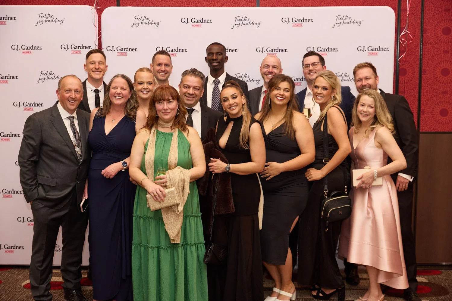 Group of men and women in formal attire posing together at a celebration or event, standing in front of a backdrop with G.J. Gardner HOMES logos and slogan.