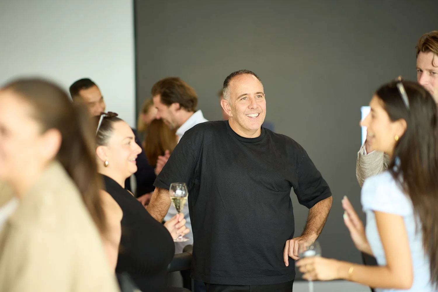 Group of people at a social event, engaging in conversation and smiling, with one man in the center holding a glass of wine.